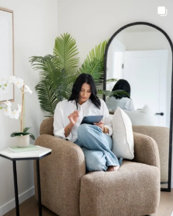 Woman sitting on a beige armchair, using a tablet, in a room decorated with large green plants, a mirror, and a side table with a white orchid plant.
