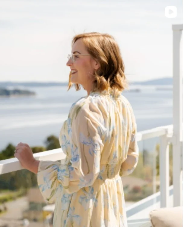 A woman with red hair, glasses, and a smile, standing on a balcony overlooking a body of water, with land in the distance, wearing a light-colored, patterned dress.