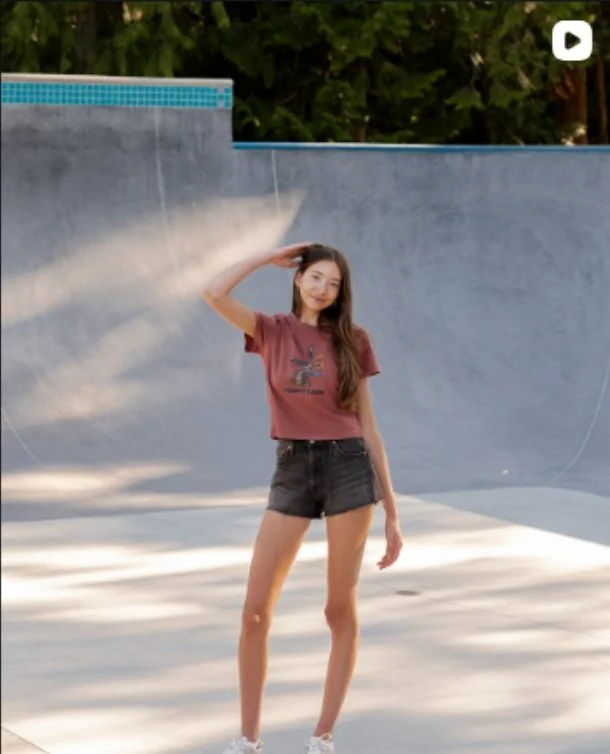 Young woman standing on skateboard at skate park bowl with gray concrete surfaces and green trees in background, wearing a maroon t-shirt and black shorts.