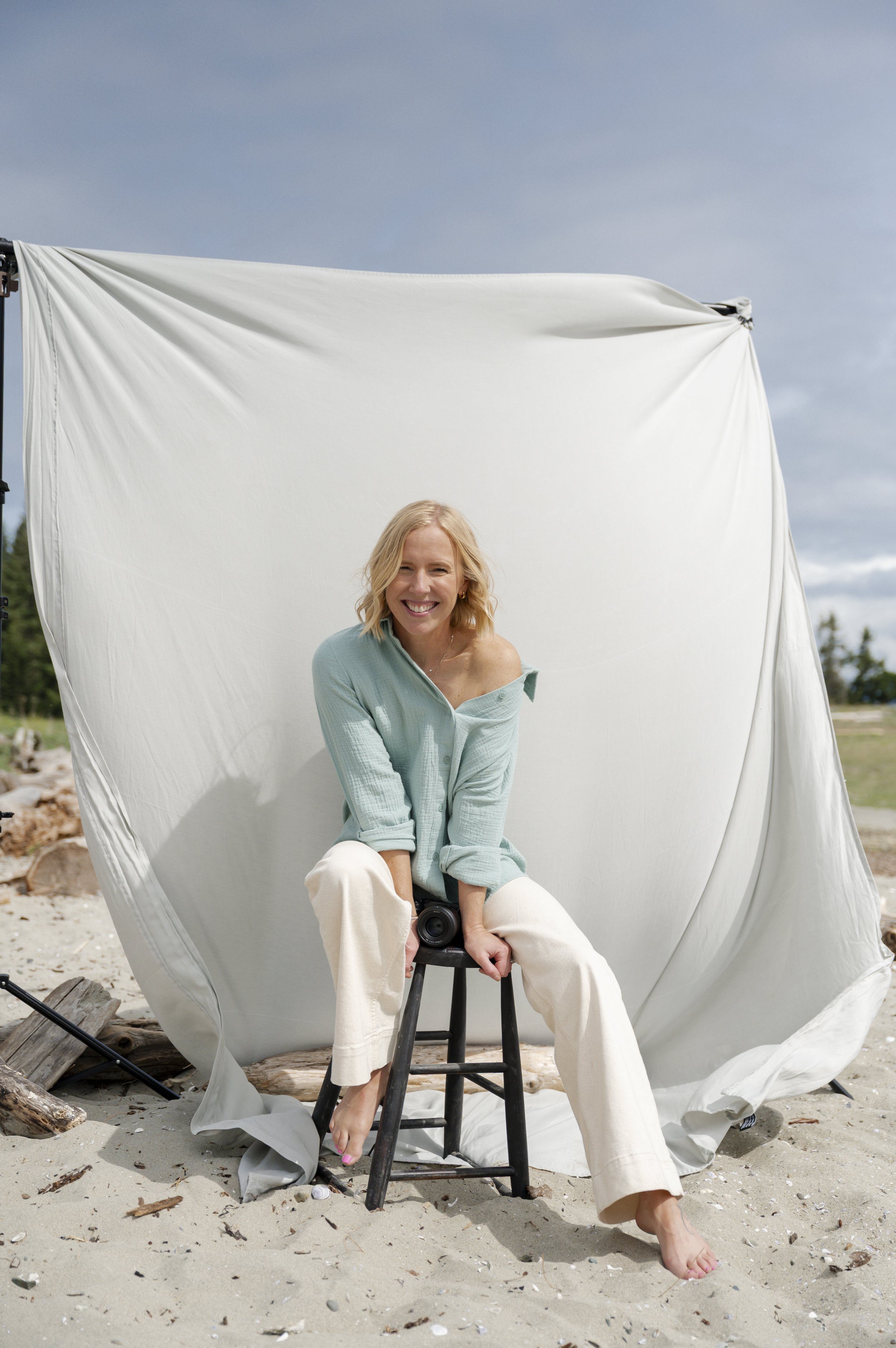 A woman smiling, sitting on a small stool on a beach, holding a camera, with a large white fabric backdrop behind her, and a cloudy sky above.