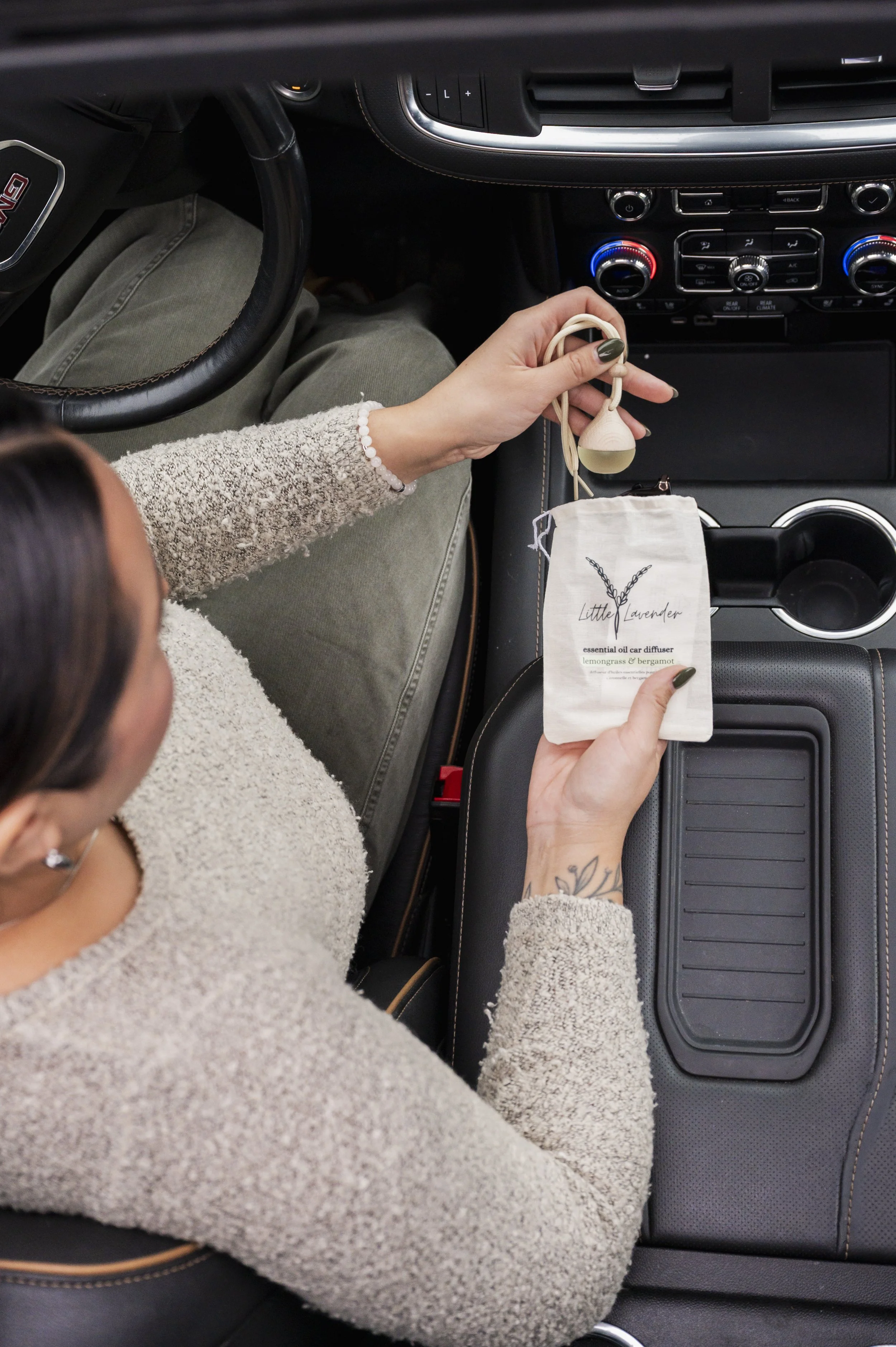 A woman sitting in a car, holding a Little Lavender essential oil car diffuser with a bottle and a cloth bag.