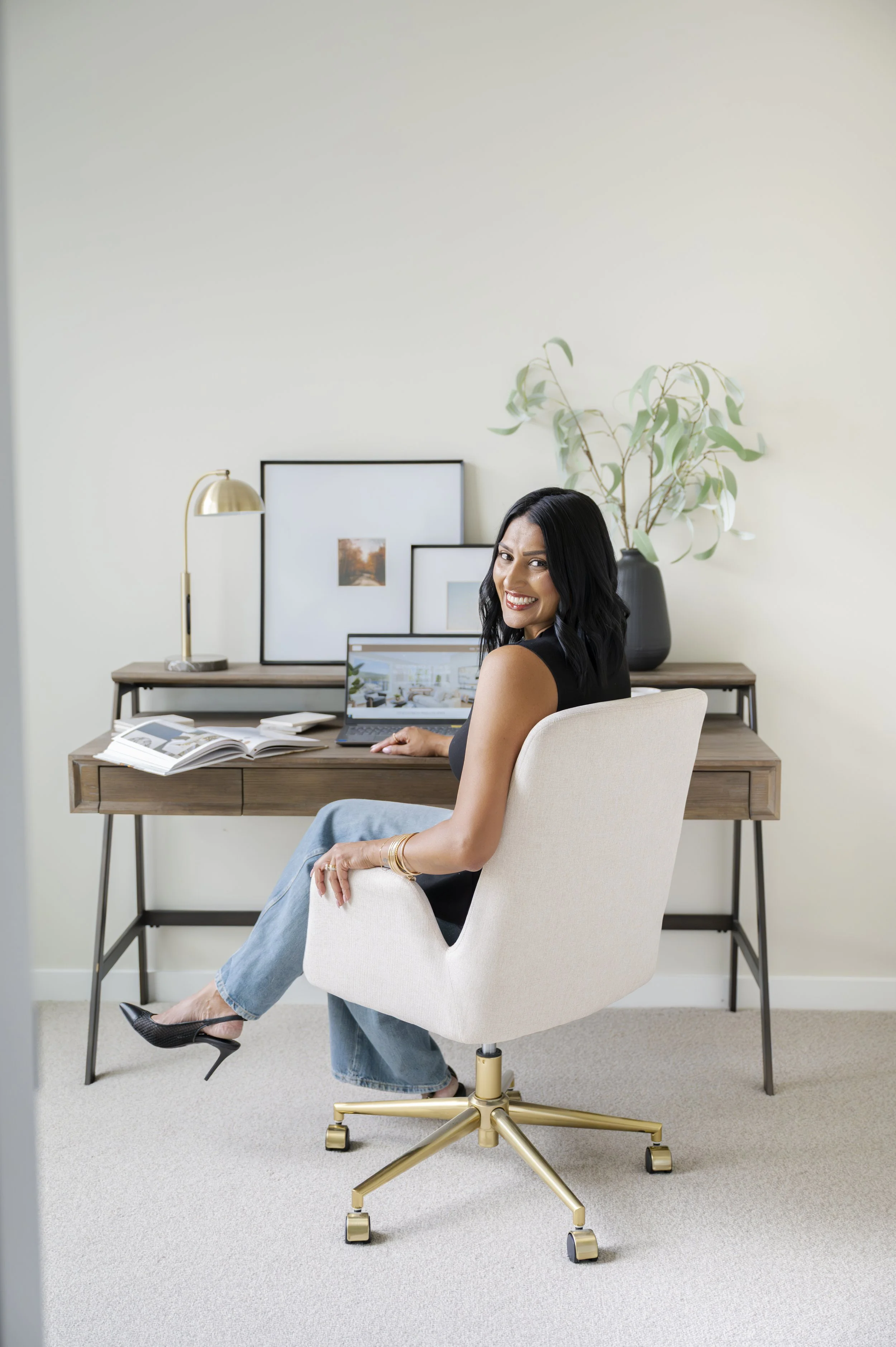 A smiling woman with black hair, wearing a sleeveless black top and light blue jeans, sitting in a white office chair in a home office. She is sitting sideways in front of a wooden desk with open magazines, a laptop, and a framed picture, with her right arm resting on the chair's armrest. Behind her is a desk with framed artwork, a modern gold desk lamp, and a large black vase with tall green leaves. The room has a light color scheme with a neutral carpeted floor.