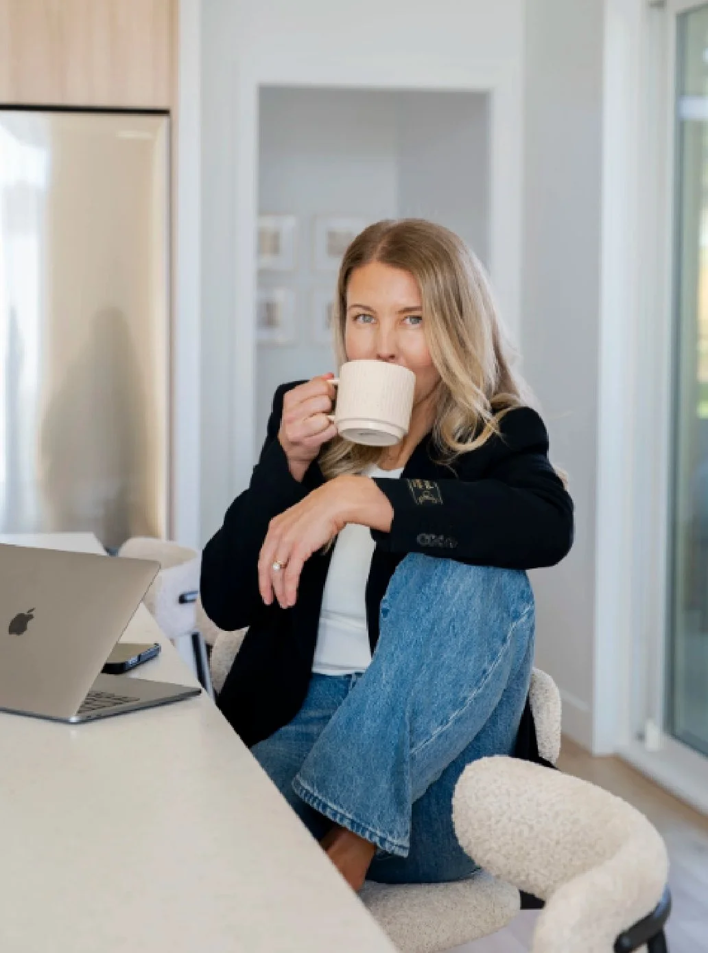A woman sitting at a table with a laptop, holding a mug, in a bright kitchen with a window on the right.