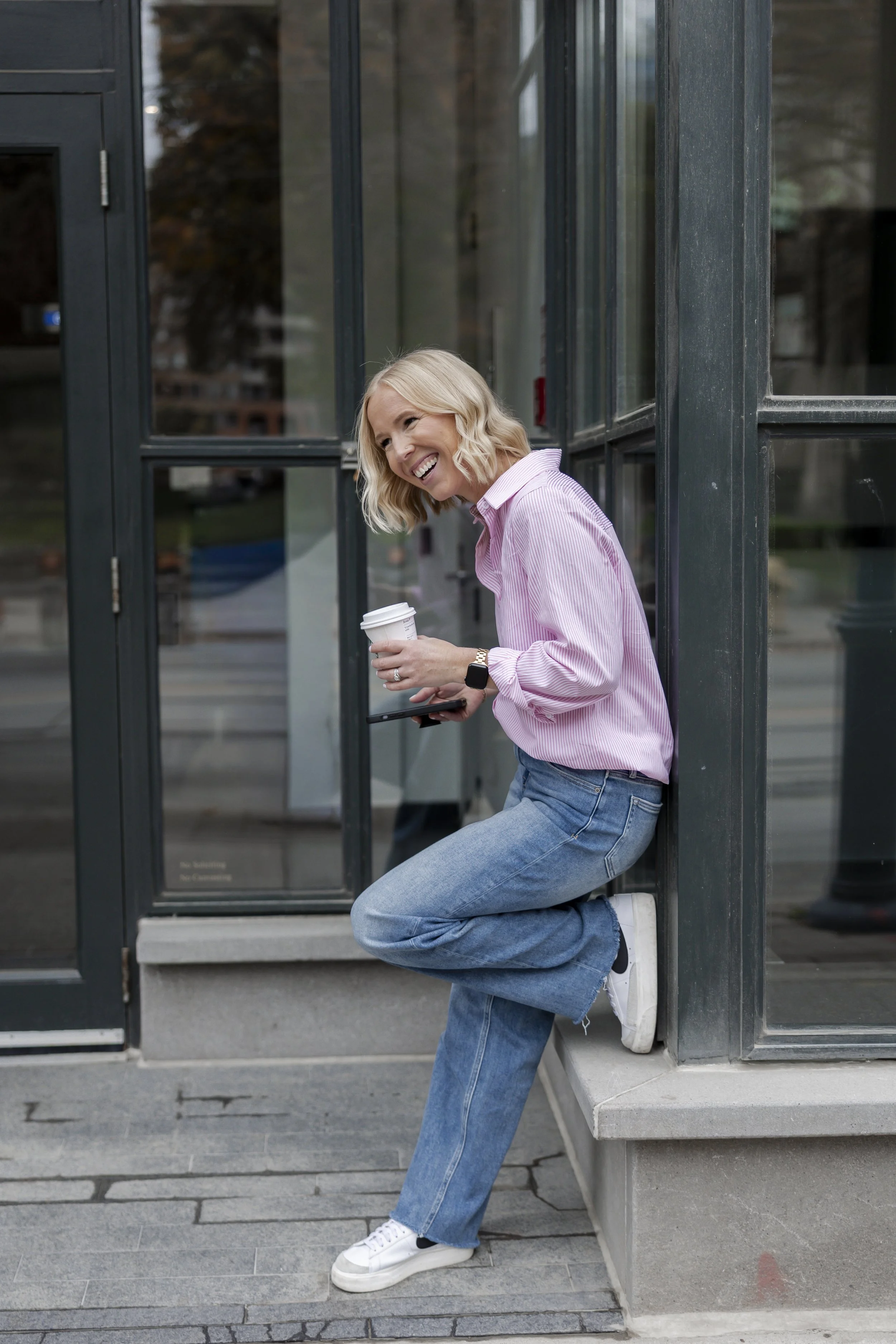 A woman with short blonde hair wearing a pink striped shirt, blue jeans, and white sneakers is standing outside a glass door, holding a coffee cup and smiling.