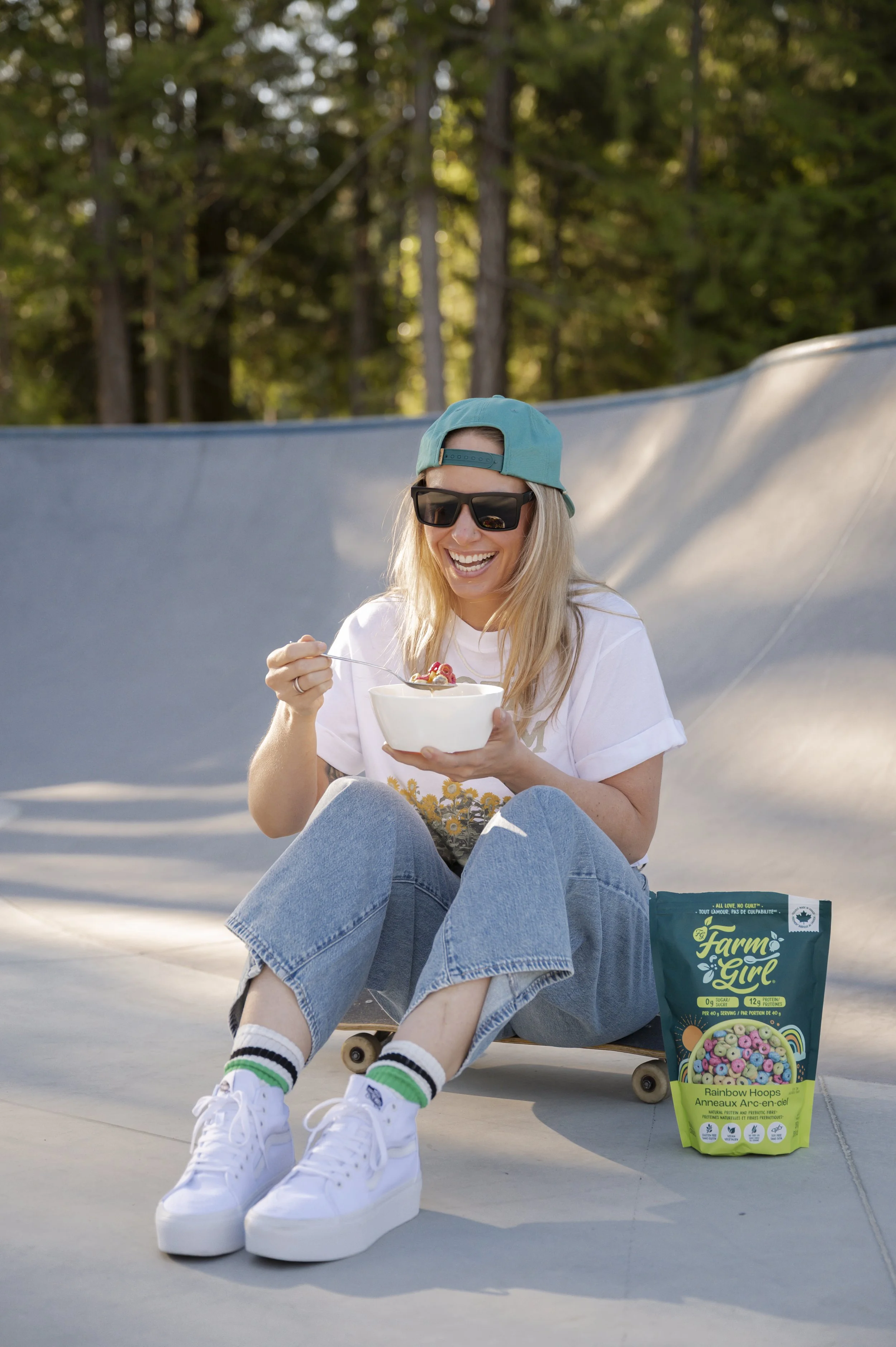 A young woman with blonde hair wearing sunglasses, a blue backward cap, a white t-shirt, and ripped jeans sitting on a skateboard at an outdoor skate park, smiling and eating cereal from a white bowl. A bag of rainbow-colored cereal is placed on the ground next to her.