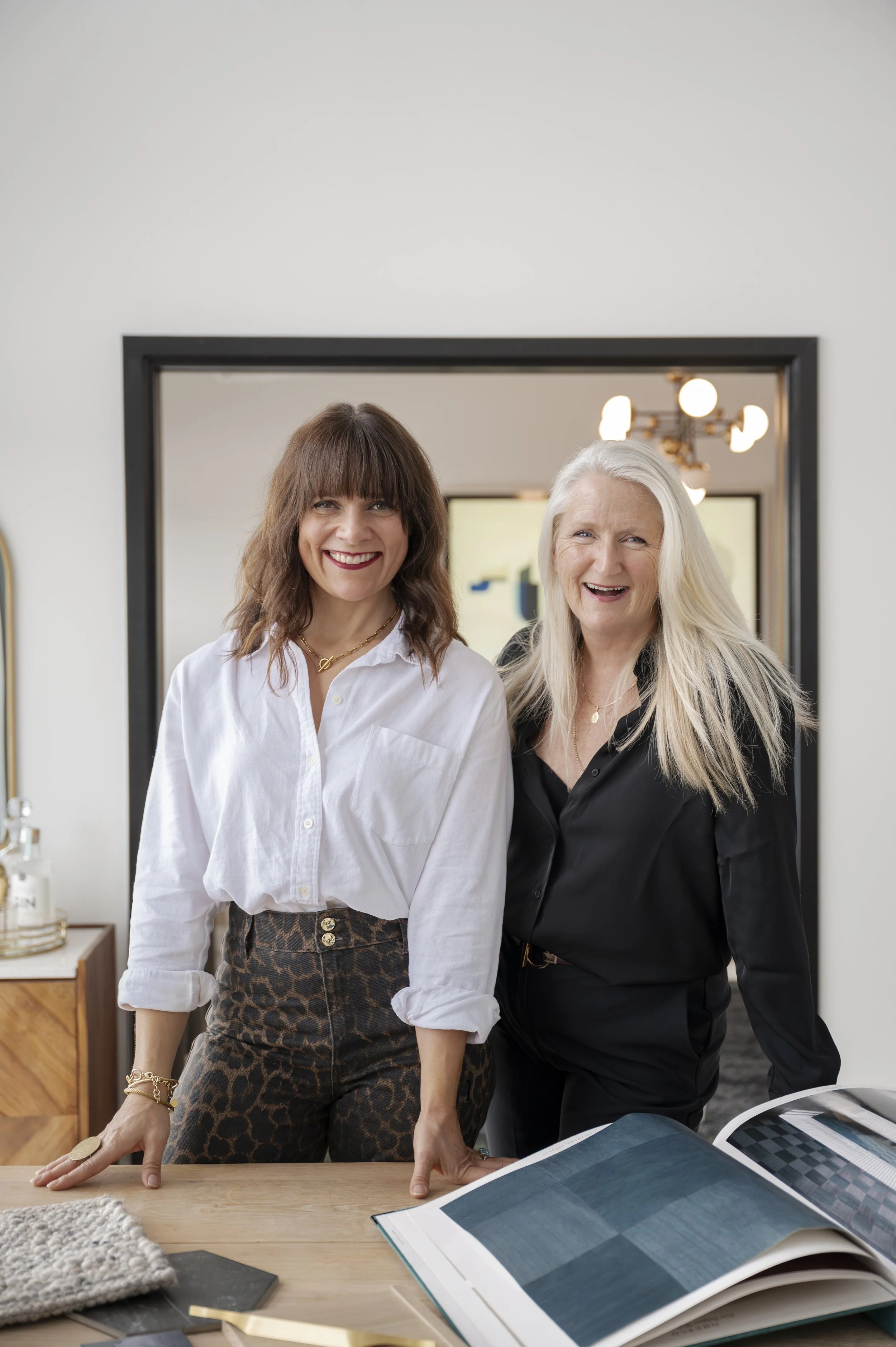 Two women in a bright modern room, smiling and leaning on a table with open design magazines, a fabric sample, and a laptop.