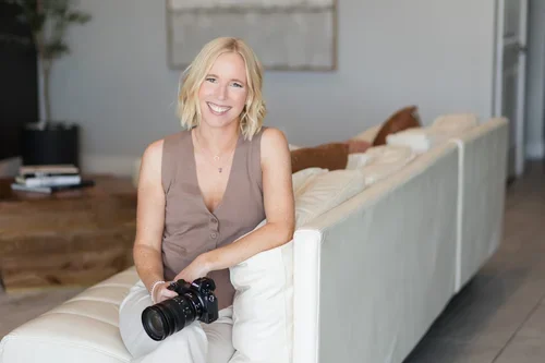 A woman with blonde hair holding a camera sitting on a cream-colored sofa in a modern living room.