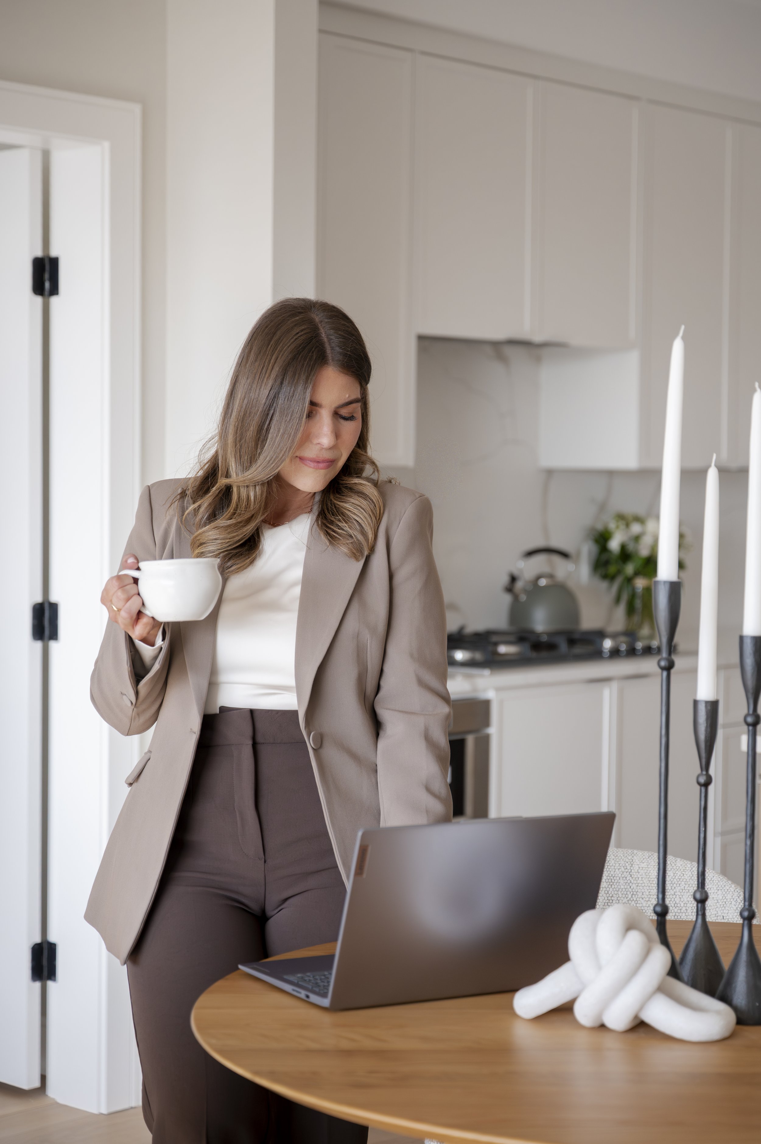 A woman with long brown hair wearing a beige blazer and brown pants holding a white mug in a modern kitchen.
