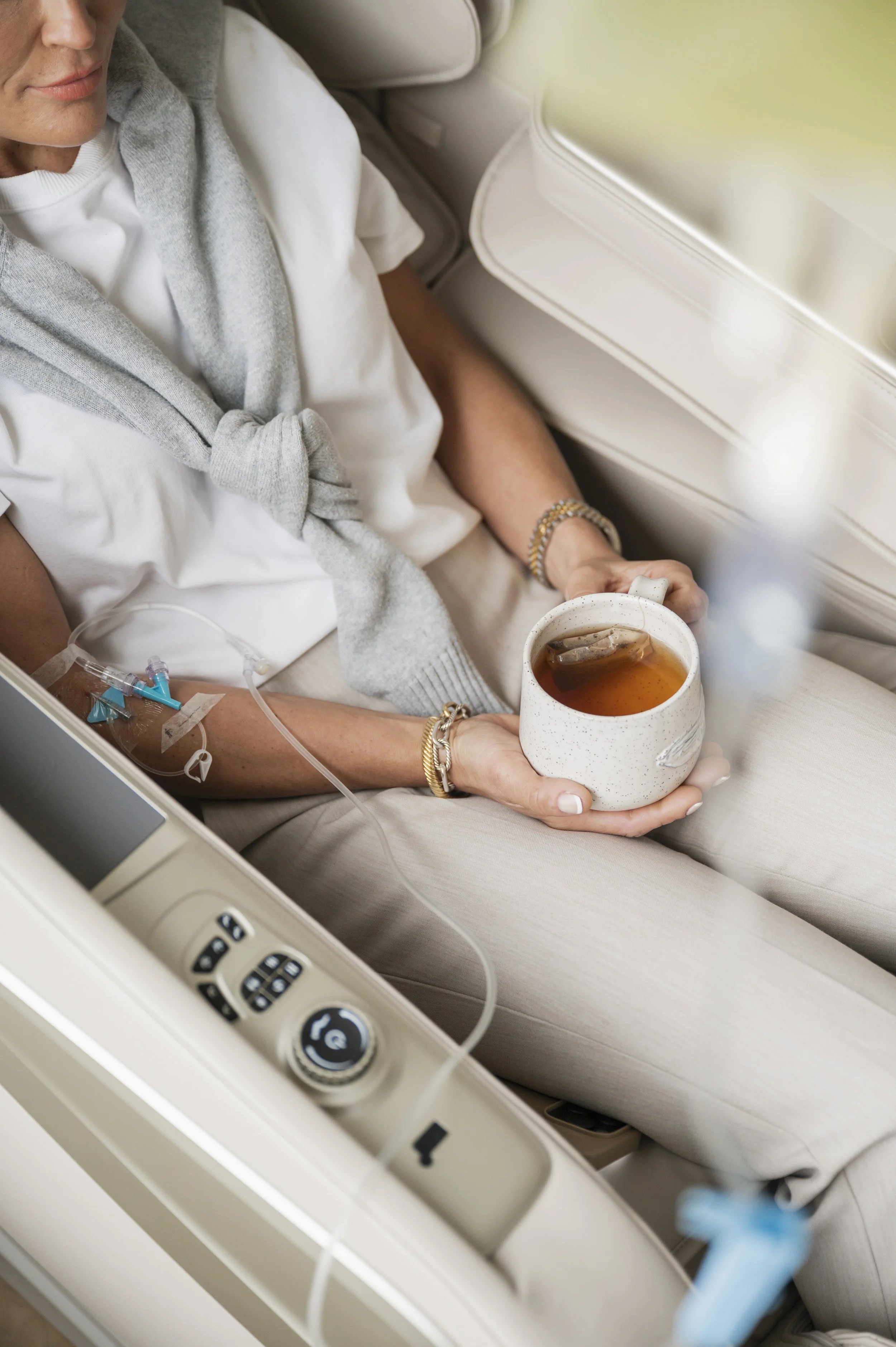 A person lies in a hospital bed with an IV in their arm, holding a mug of tea, with medical equipment and control panel visible nearby.