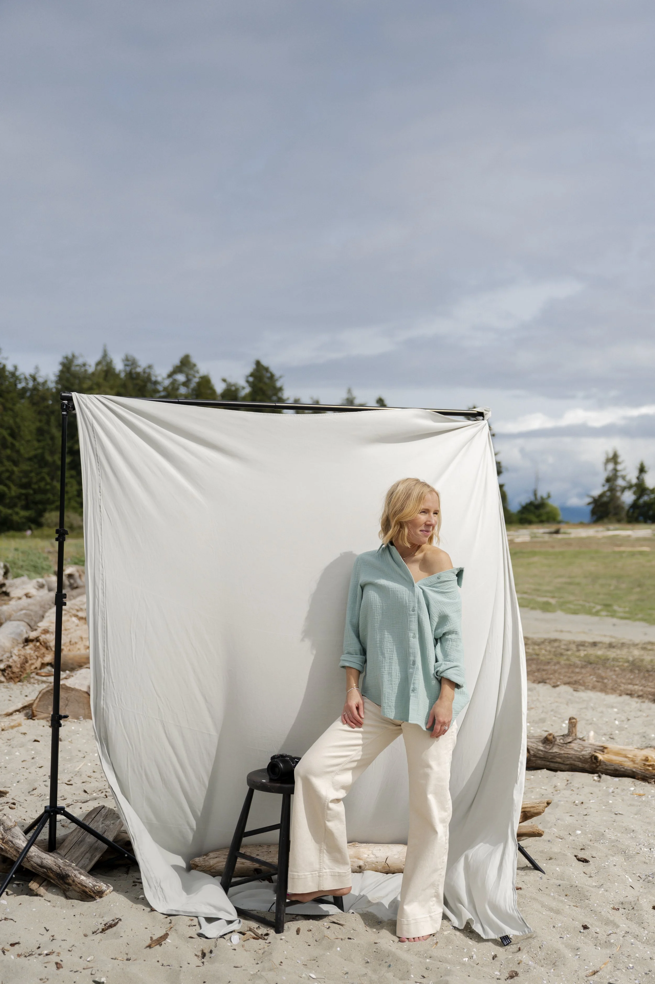 A woman with blonde hair standing on a sandy beach, partially wrapped in a light blue shirt, posed in front of a white backdrop with trees and cloudy sky in the background.