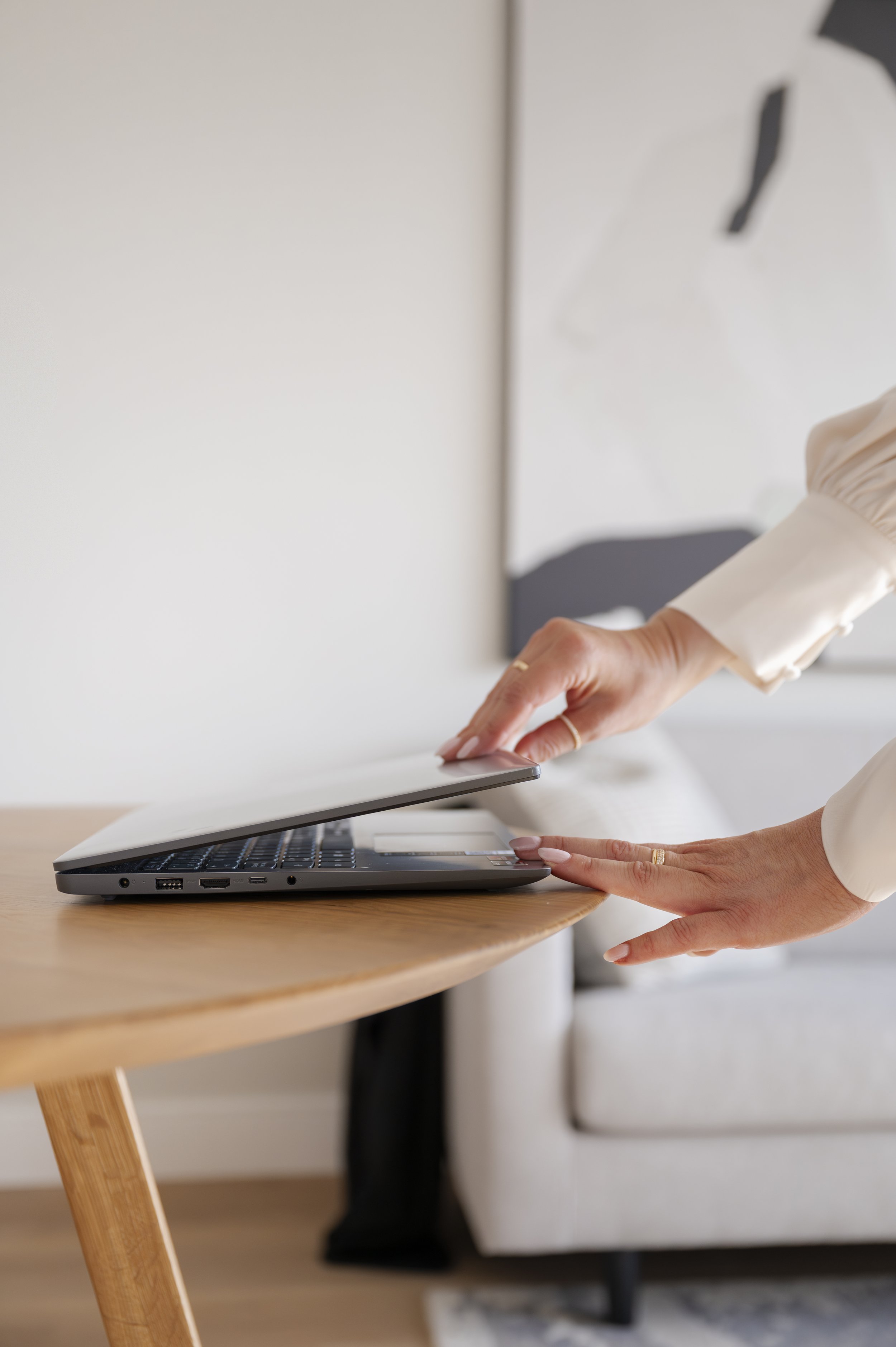 Person using a laptop at a wooden desk, lifting the screen open with one hand, in a bright room.