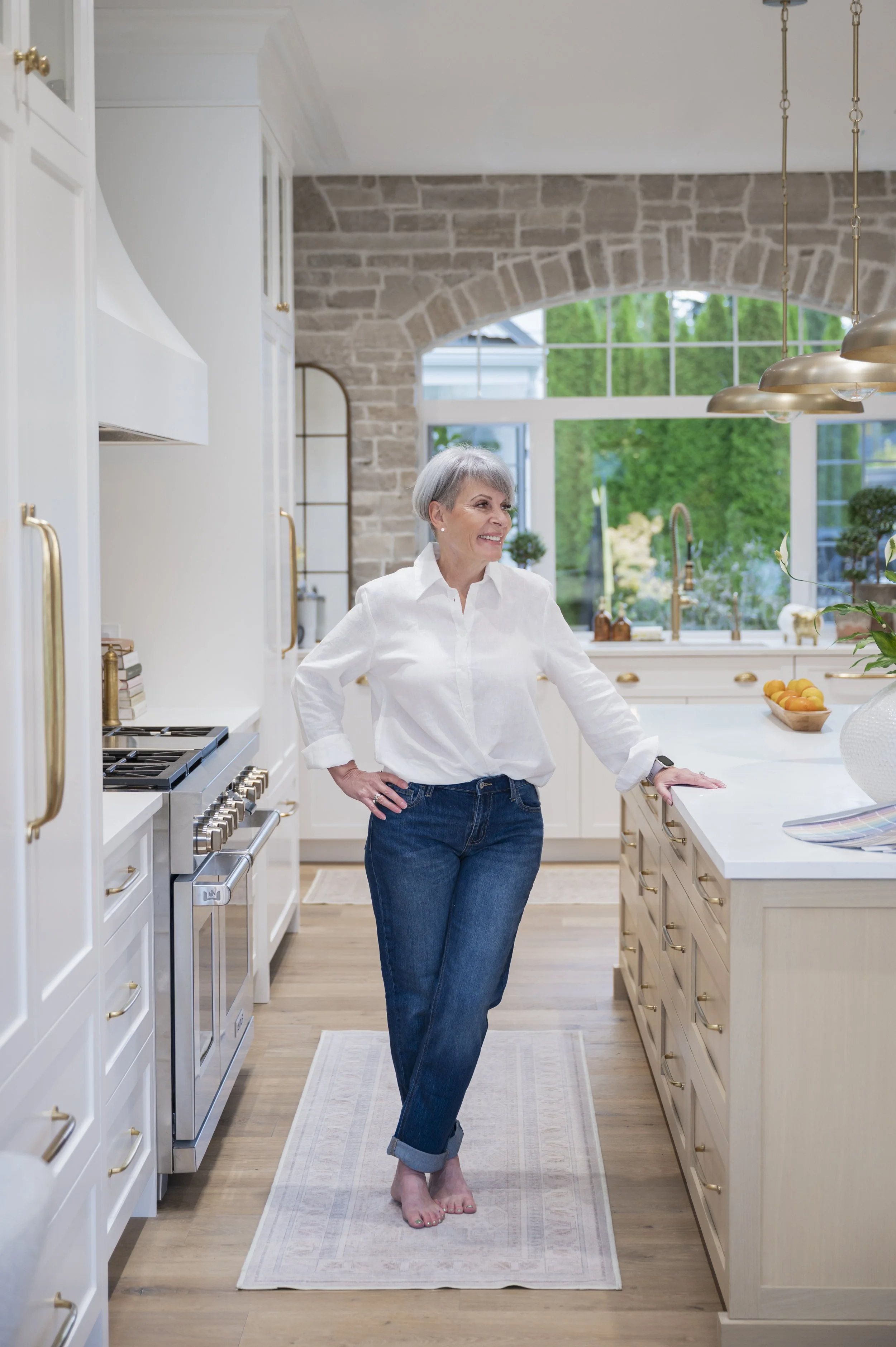 A woman with short gray hair, wearing a white shirt and blue jeans, stands barefoot in a bright, modern kitchen with white and wooden cabinets, a large window with a green view, and gold accents. She is smiling and leaning on the kitchen island.