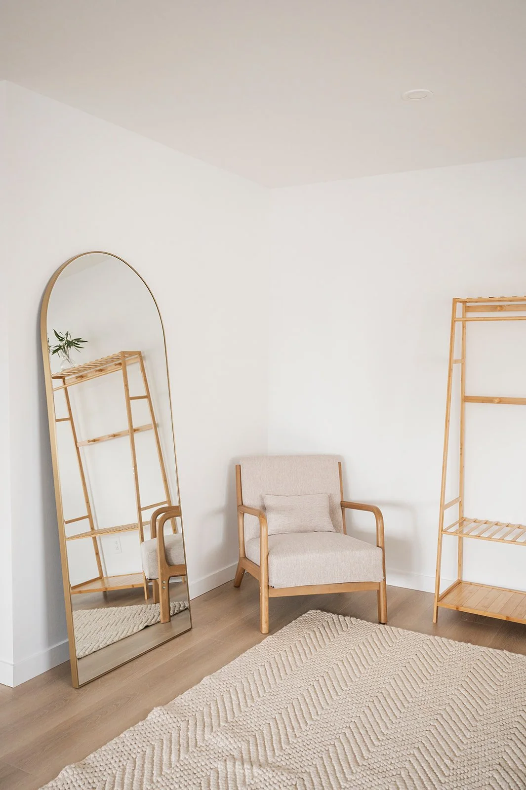 Living room with a beige armchair, a tall oval mirror leaning against the wall, a wooden shelf, and a cream-colored textured rug on light wood flooring.