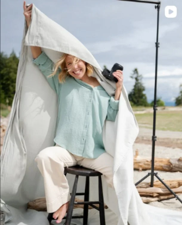 A woman sitting on a stool outdoors, smiling, holding a camera, with a large white sheet draped over her head and holding it up, set up as a makeshift backdrop for a photo shoot.