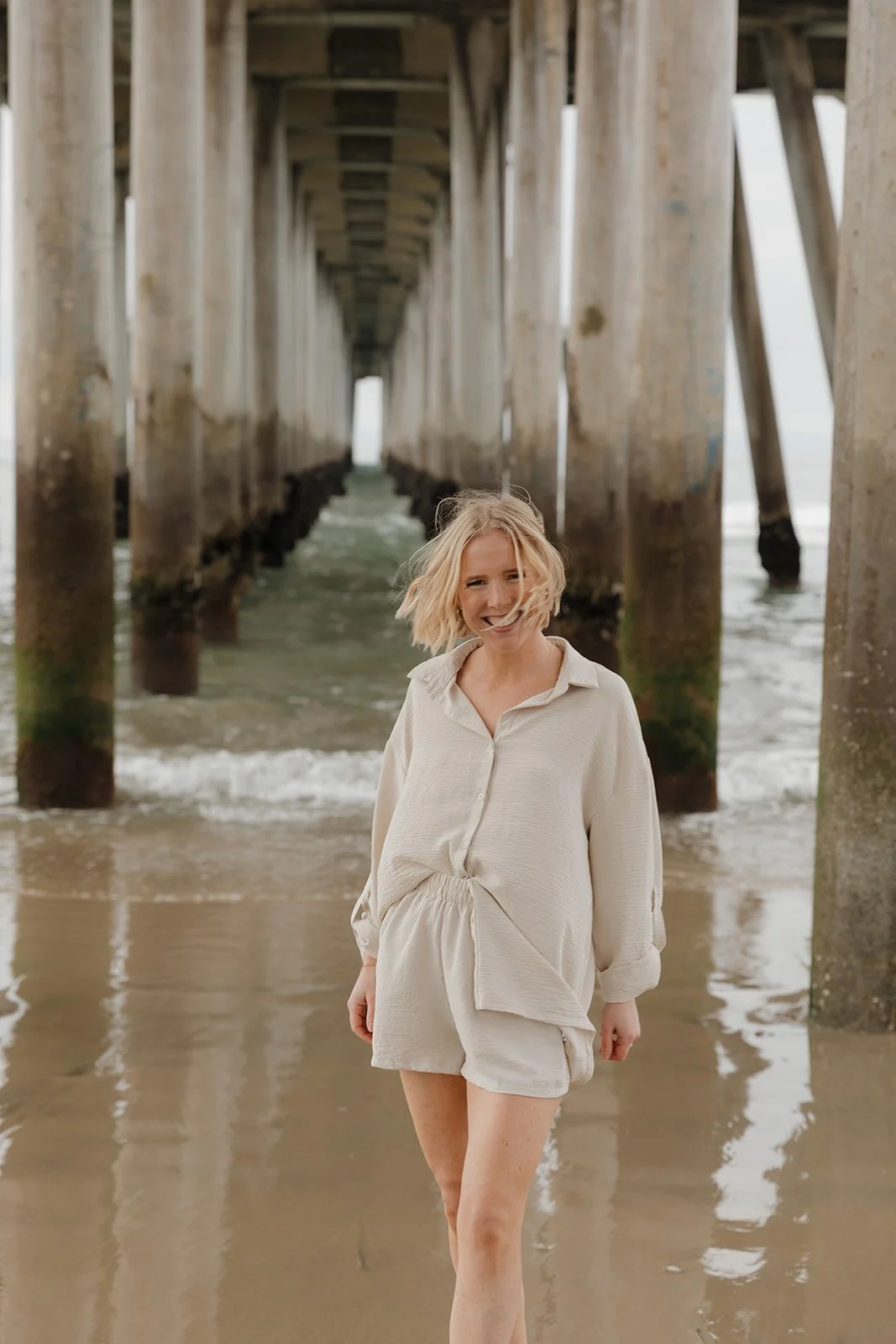 A woman stands on the beach under a pier, smiling, with ocean water around her legs and wooden pier supports in the background.