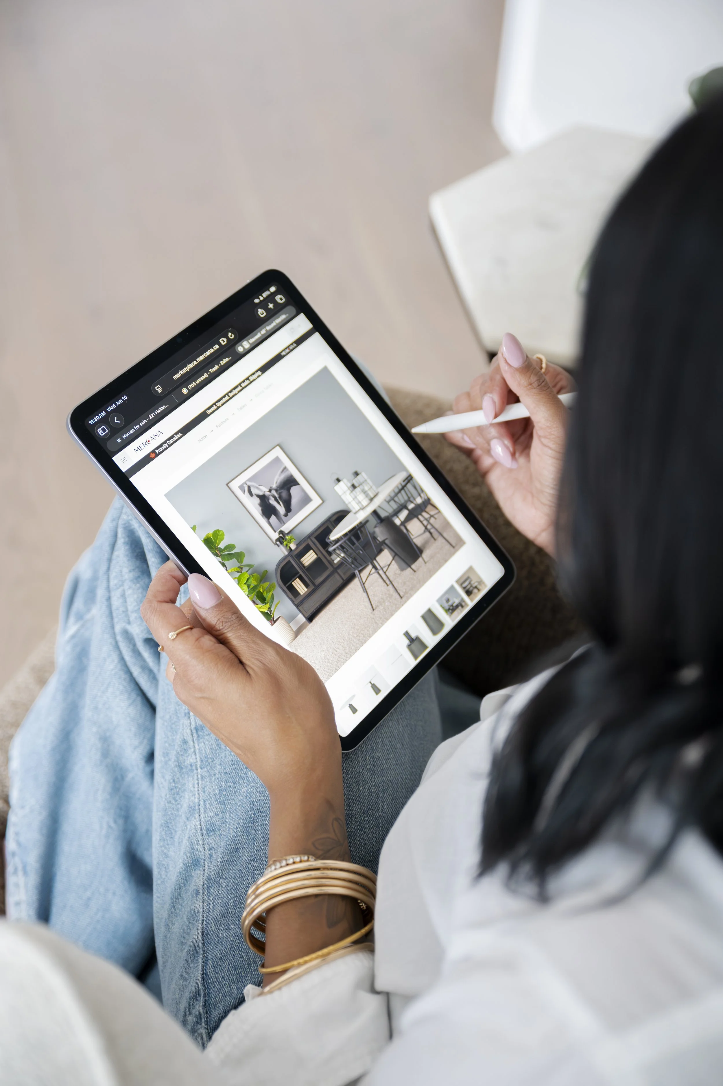 A person holding a tablet with an interior design website open, seated with crossed legs wearing a white shirt and gold bangles. The person is using a stylus on the tablet, which displays a room with furniture and decor.