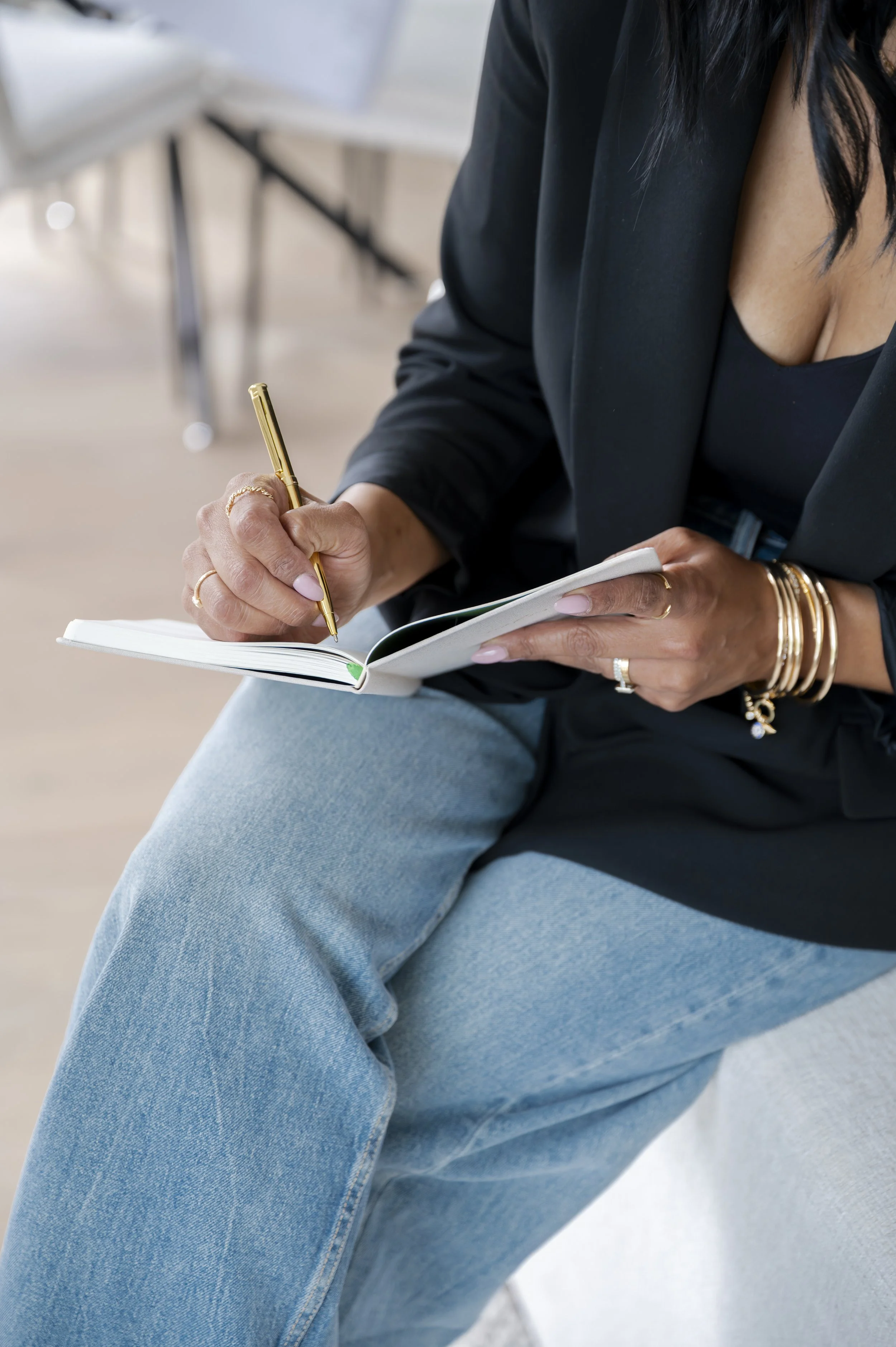 A woman wearing a black blazer and light blue jeans sits on a chair, writing in a notebook with a gold pen. She has multiple gold bangles and rings, and her black hair is visible. The background is blurred with a light and neutral color palette.