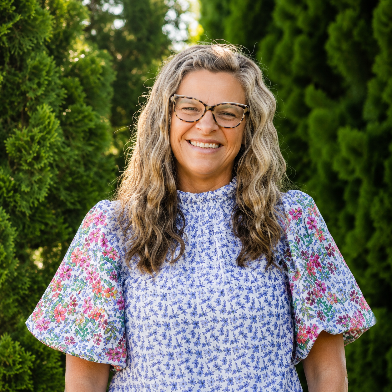 A woman with long, wavy hair wearing glasses and a floral-patterned dress, smiling outdoors with green trees in the background.