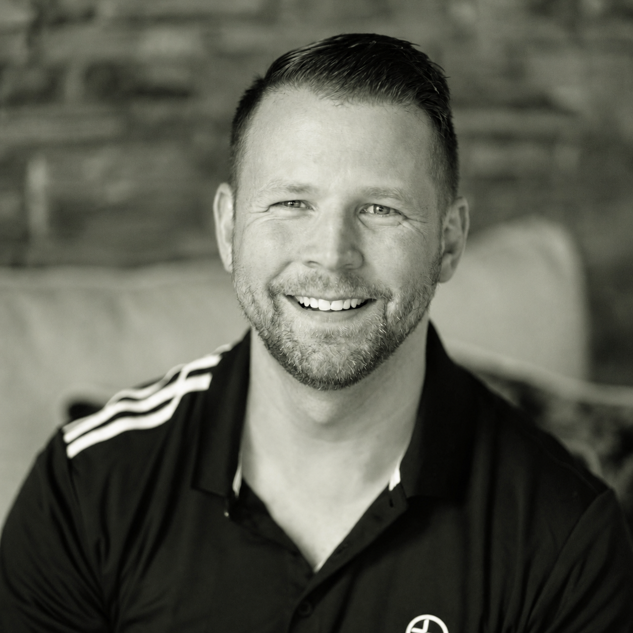 A smiling man with short hair and a beard, wearing a black polo shirt with white stripes on the shoulders, sitting indoors with a brick wall in the background.