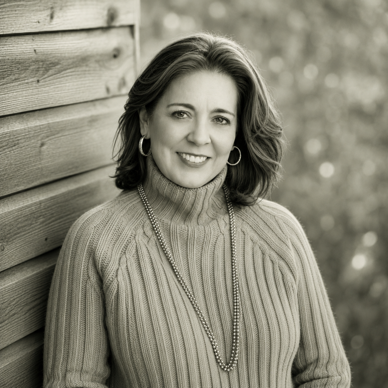A woman with shoulder-length hair, wearing a turtleneck sweater and jewelry, is smiling outdoors against a wooden wall with blurred background.