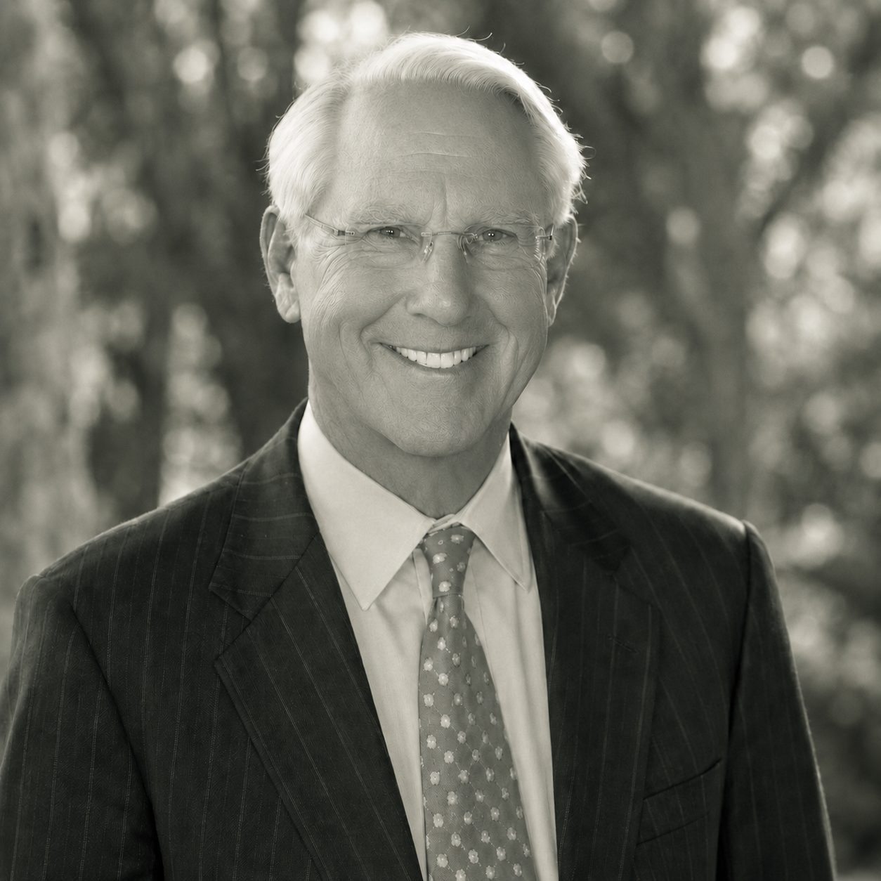 A smiling elderly man with light hair and glasses, wearing a suit and tie, stands outdoors with a blurred background of trees.
