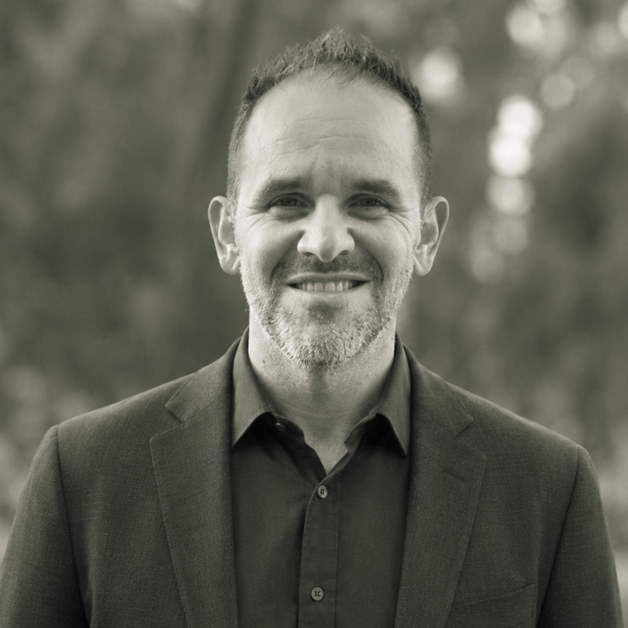 A smiling man with short hair and a beard, wearing a dark blazer and shirt, standing outdoors with a blurred natural background.