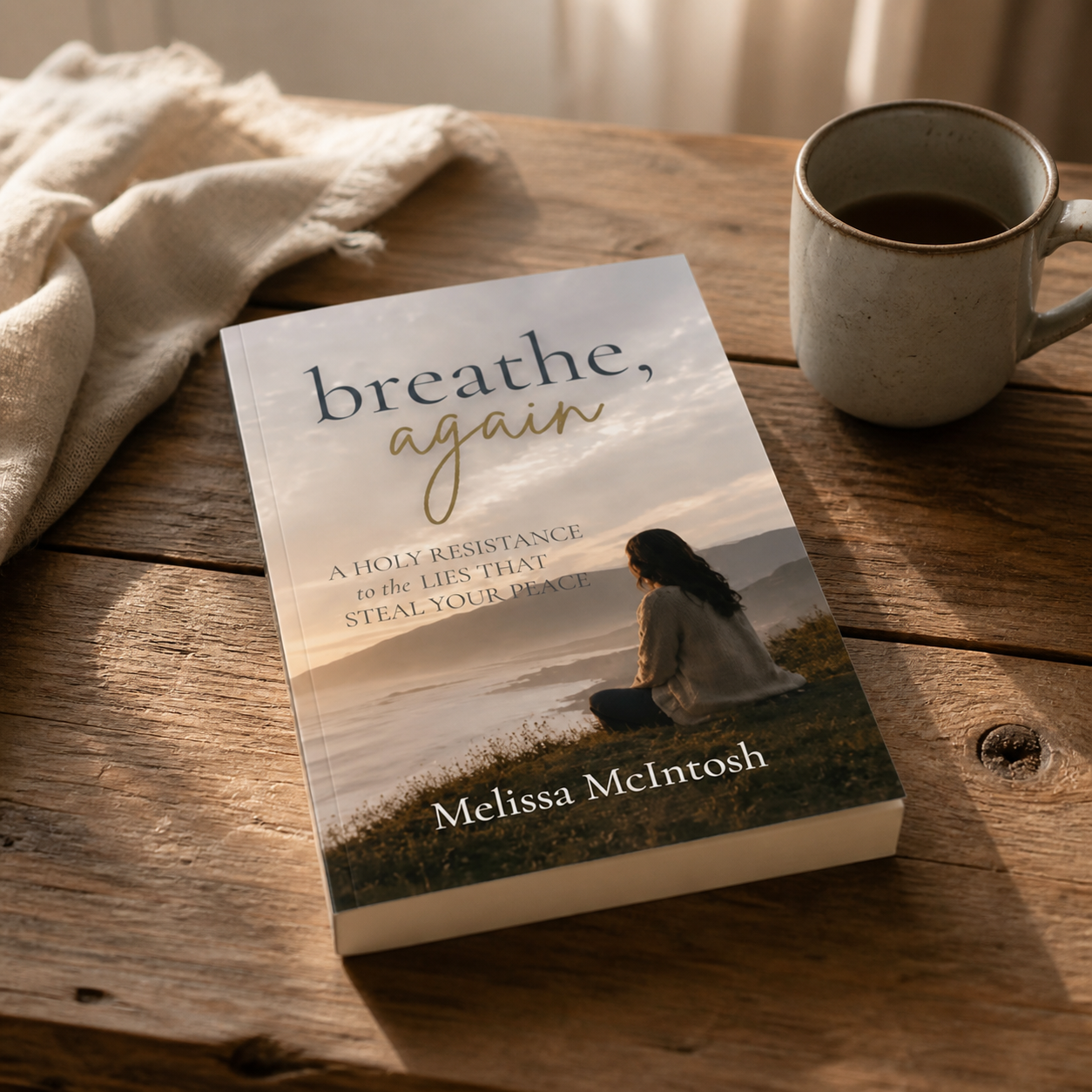 A book titled "breathe, again" by Melissa McIntosh on a wooden table next to a cup of coffee and a beige cloth.