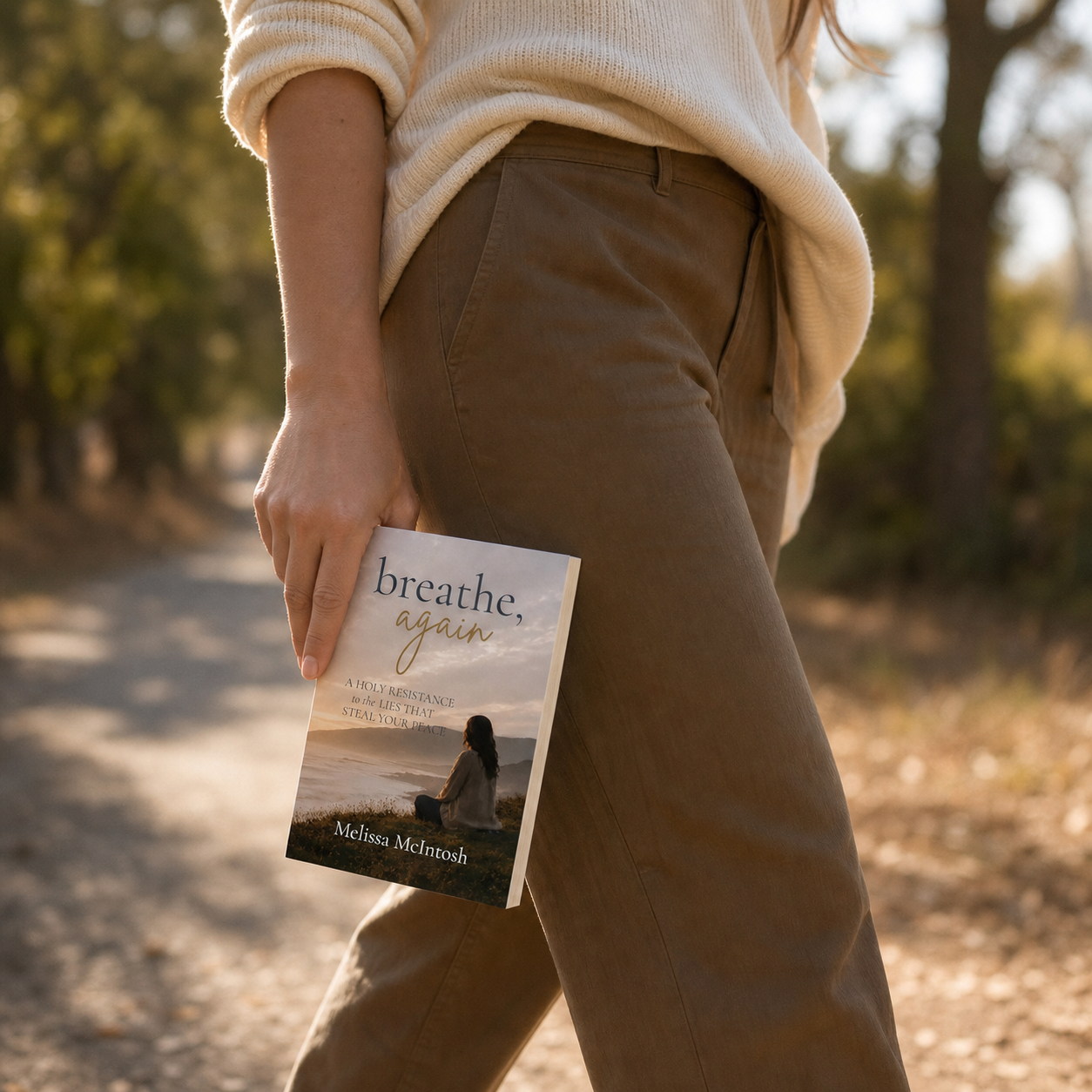 Person walking outdoors holding a book titled 'breathe, again' by Melissa McIntosh, with trees and a dirt path in the background.