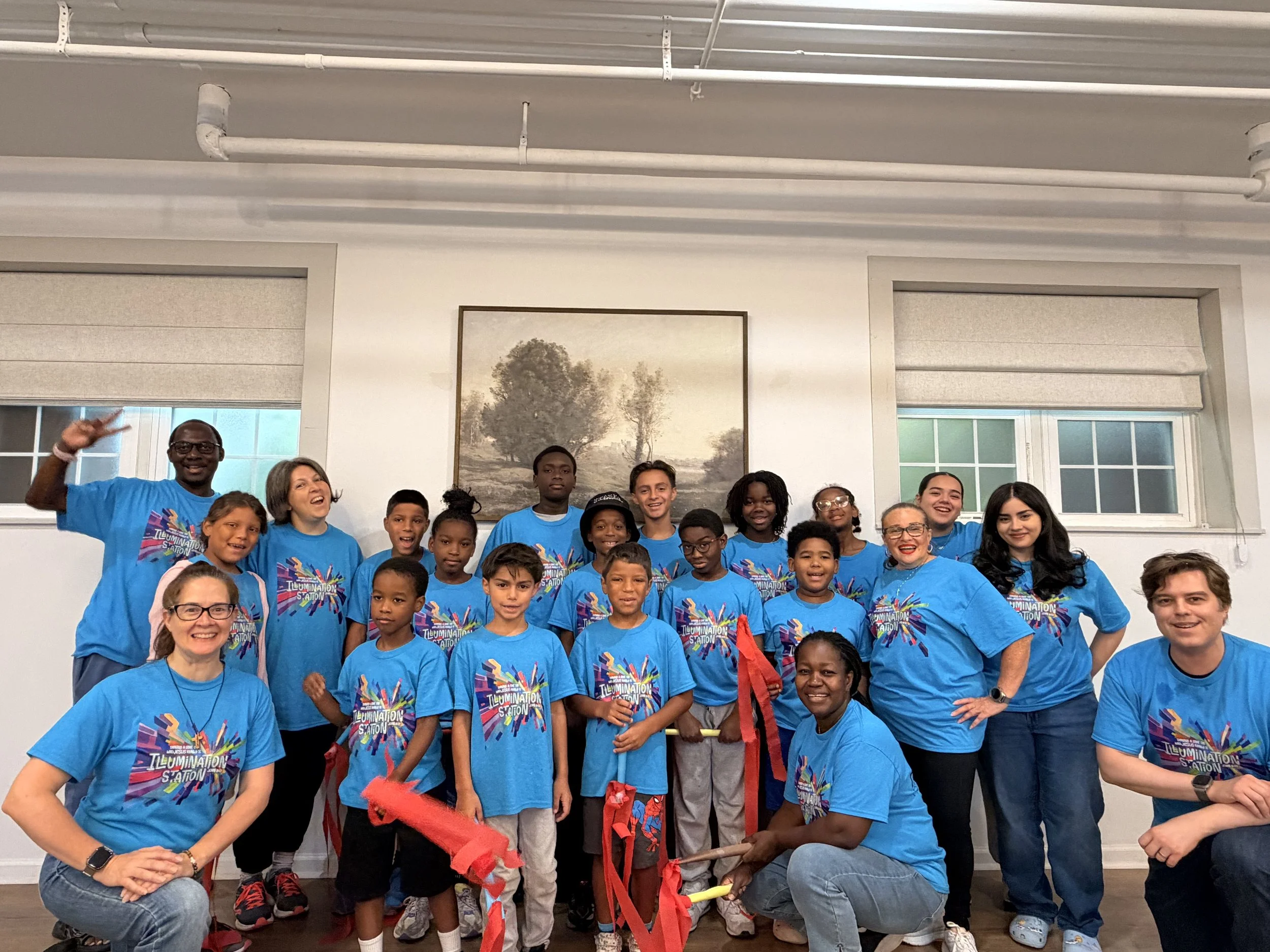 Group photo of children and adults wearing matching blue T-shirts with a colorful logo, gathered indoors in front of a landscape painting and large windows, celebrating an event.