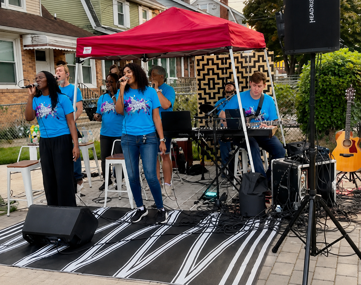 A group of young people performing music outdoors under a red canopy, with three women singing into microphones and two men playing keyboard and guitar, set up on a sidewalk with a black and white striped rug, surrounded by musical equipment and a microphone stand.