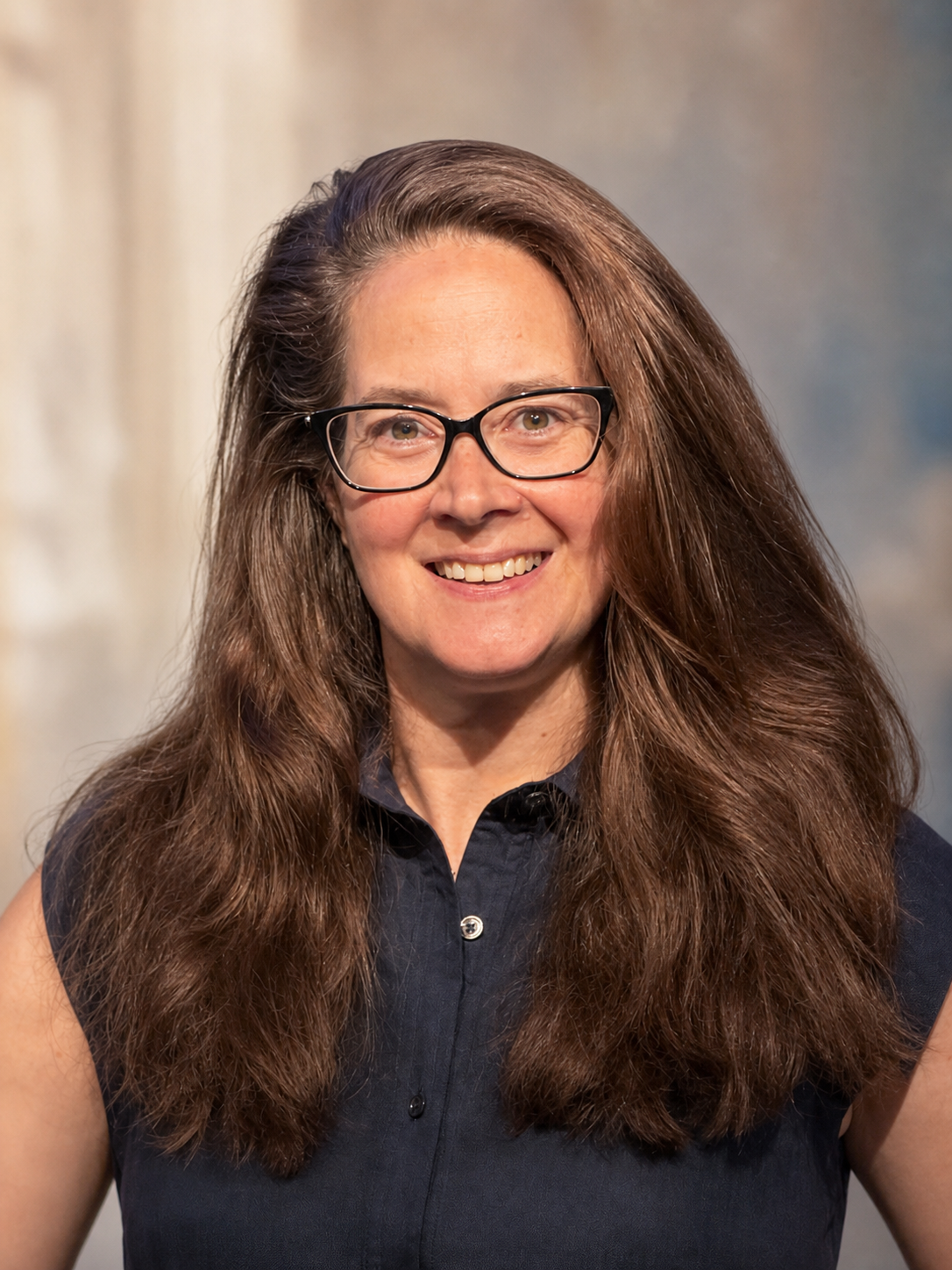 A woman with long, wavy brown hair, black glasses, and a sleeveless dark blue shirt smiling at the camera.