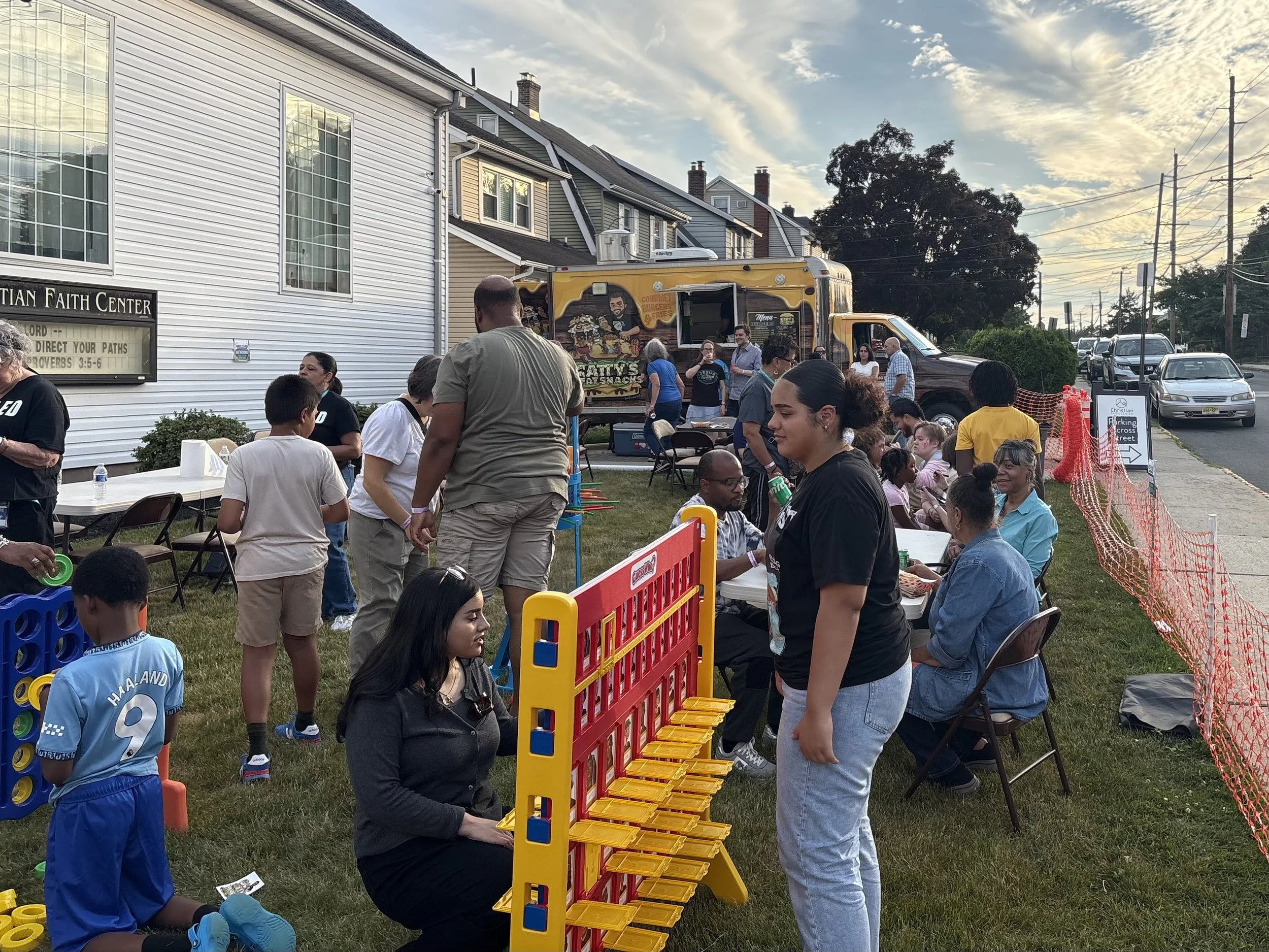 A community gathering outdoors next to a white building with a sign for a Christian faith center. People are playing giant connect four and socializing. A yellow food truck with a cartoon chef and food advertisements is parked in the background. The scene is set in the evening with a partly cloudy sky.
