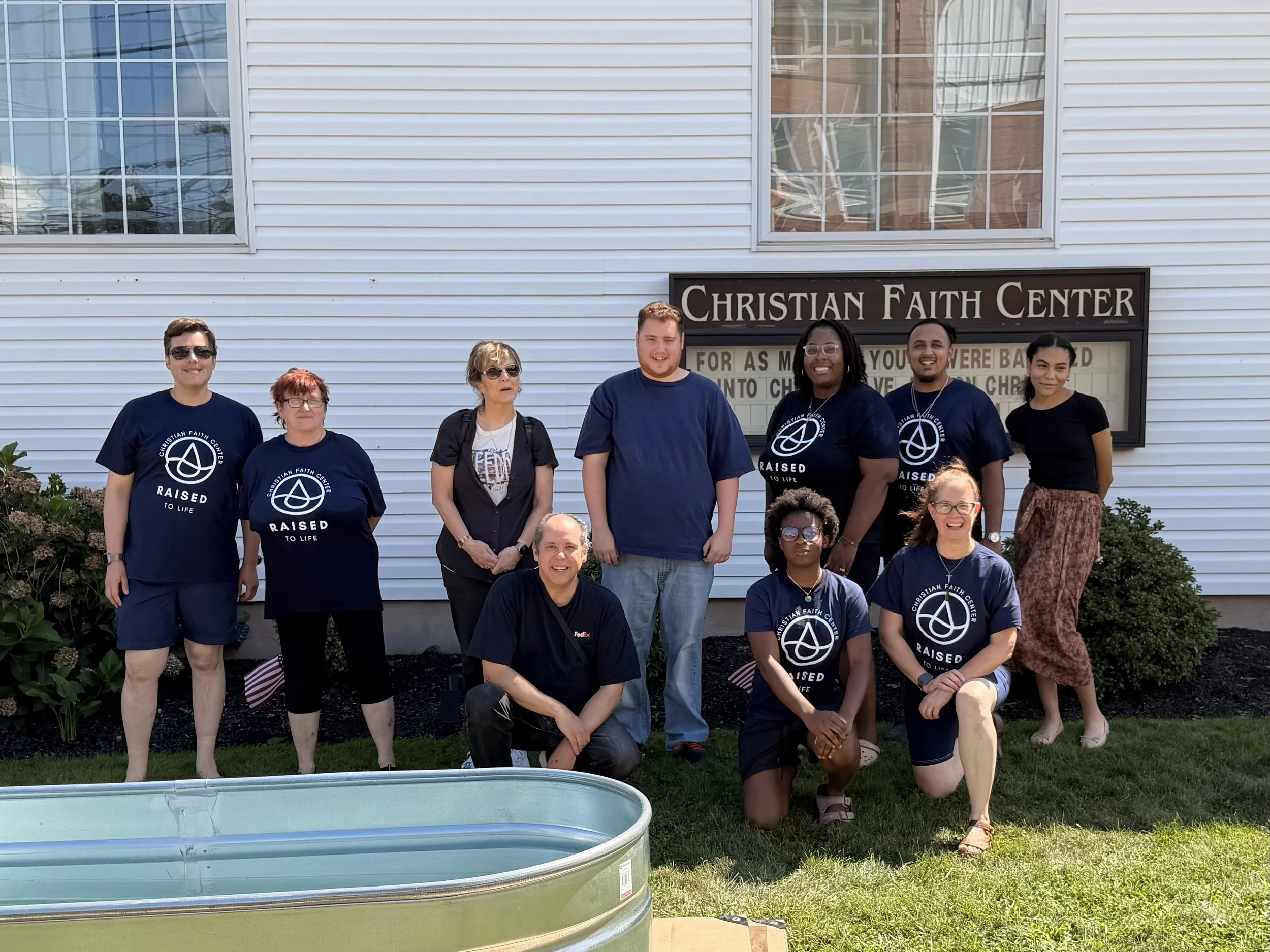 Group of people standing outside in front of a white building, some wearing matching Christian Faith Center t-shirts, posing for a photo during daytime.