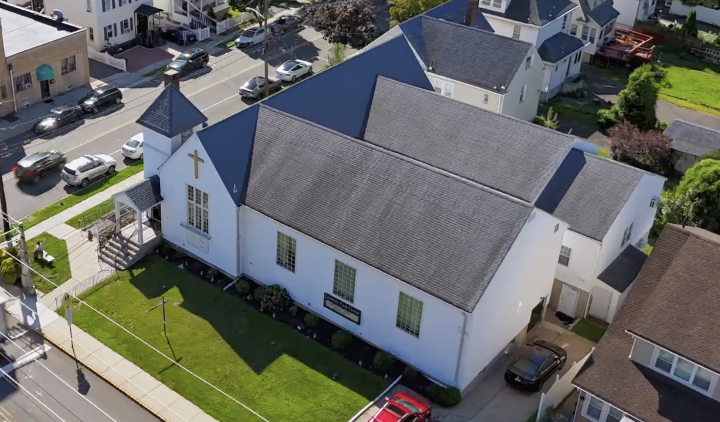 A small white church with a gray shingled roof, a gold cross on the front, and glass block windows, located on a sunny street with parked cars and neighboring houses.