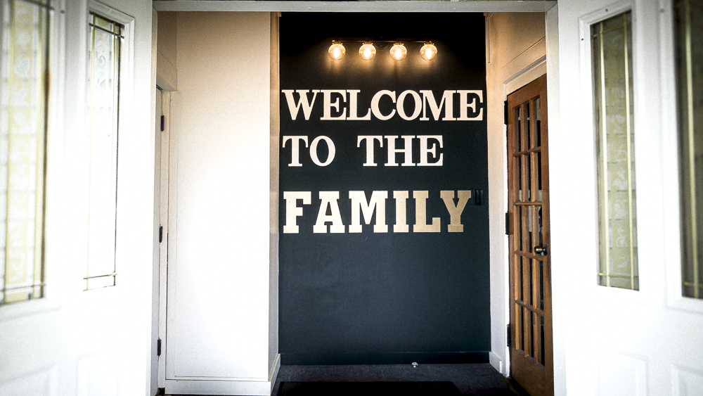 Interior view of a welcoming home entryway with a navy wall featuring large white letters spelling out "WELCOME TO THE FAMILY." Above the sign, there are five exposed light bulbs. To the right is a wooden door with glass panels, and on each side, there are white walls with large windows.