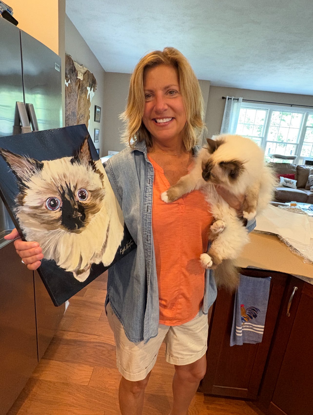 Smiling woman holding a fluffy cat beside its custom pet portrait.