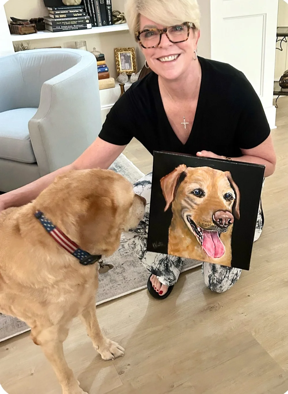A woman with blonde hair and glasses smiles while kneeling on the floor, holding a painting of a happy golden retriever with its tongue out. Next to her, a golden retriever with a patriotic collar looks at the painting in a cozy living room.