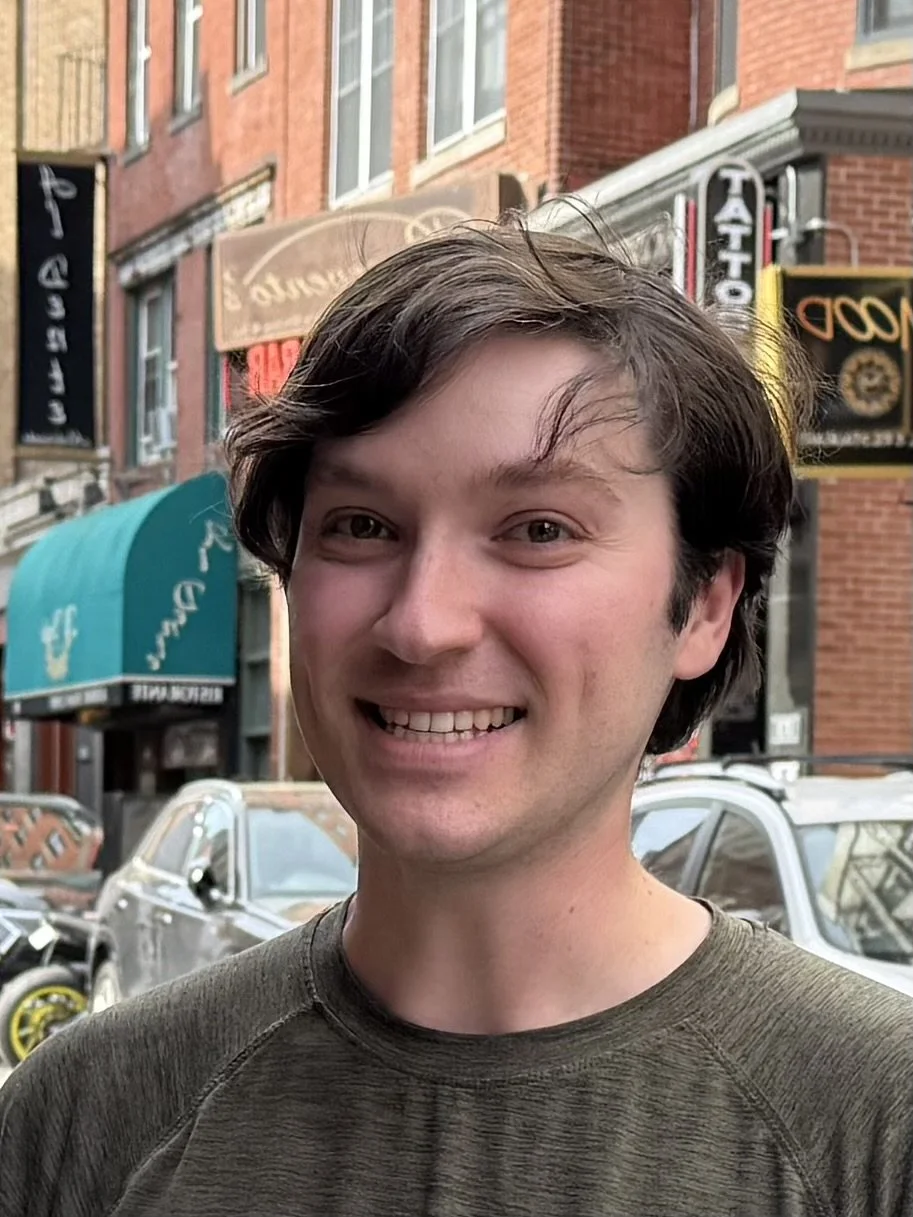 A young man with dark brown hair smiling outdoors on a city street, with cars and storefront signs in the background.