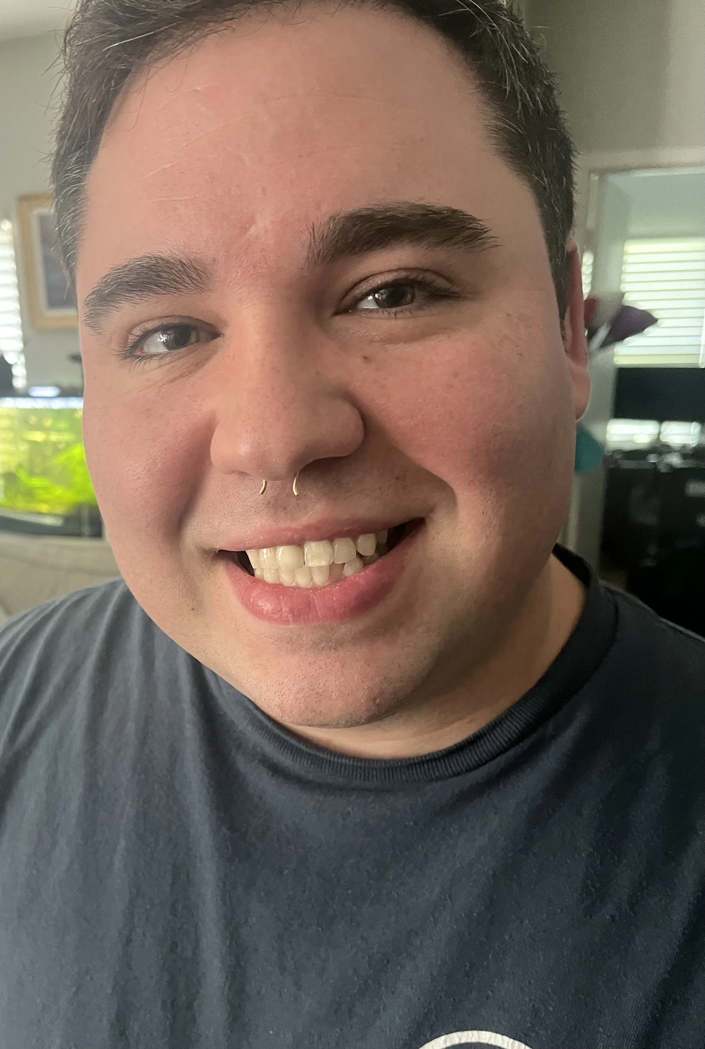 Close-up of a smiling person with short dark hair and a septum piercing, wearing a dark gray shirt. Background includes a fish tank and a window with blinds.