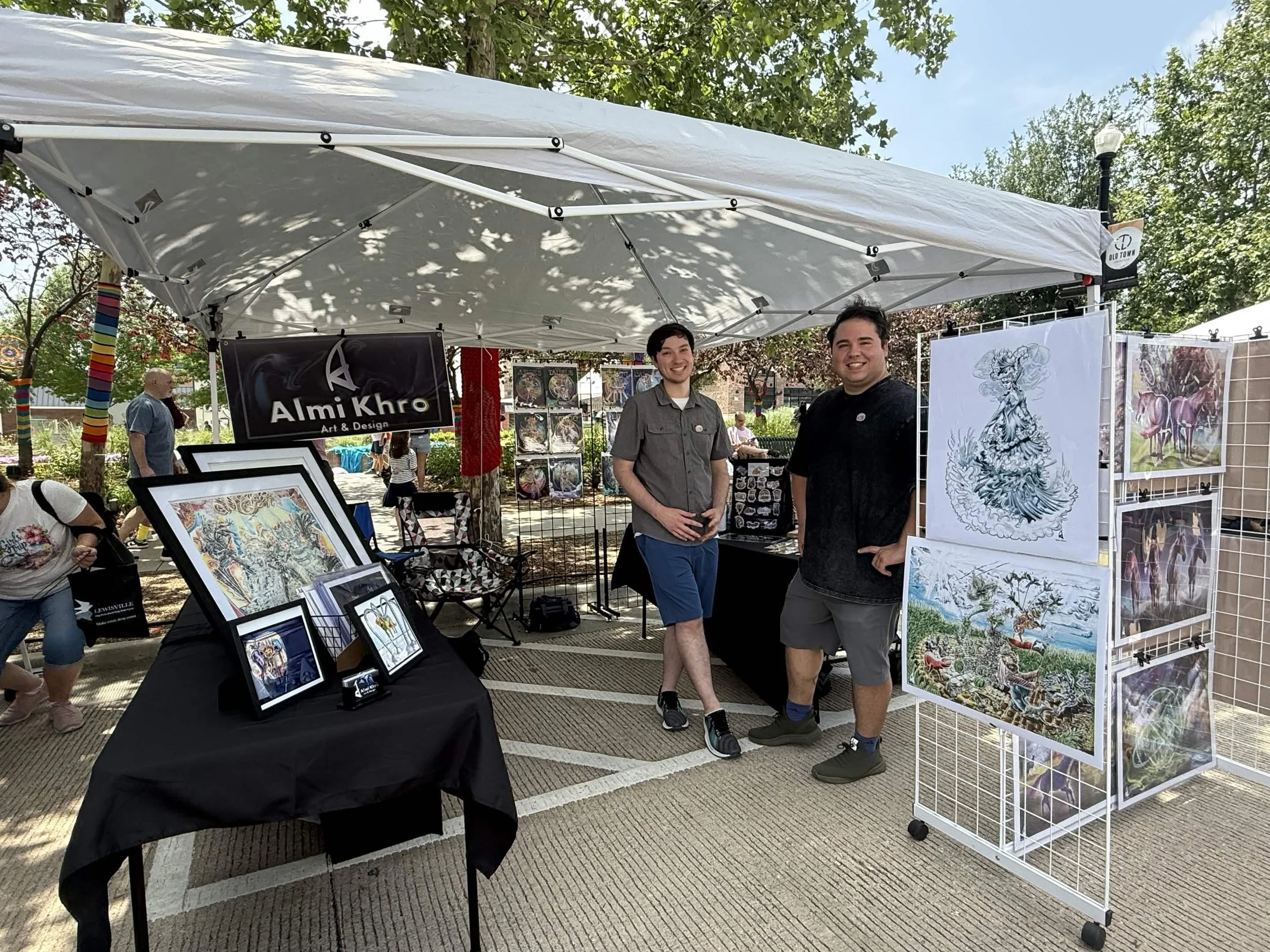 At an outdoor art fair, two young men stand under a white canopy, smiling at the camera. The booth features framed artwork and paintings on display, with trees and park elements in the background.