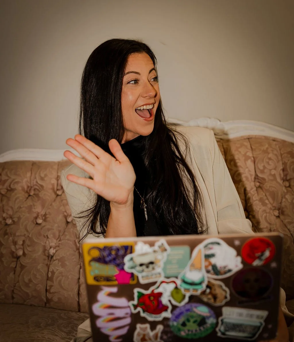 A woman with long dark hair, smiling and waving, sitting on a vintage floral-patterned couch, with a laptop decorated with colorful stickers in front of her.