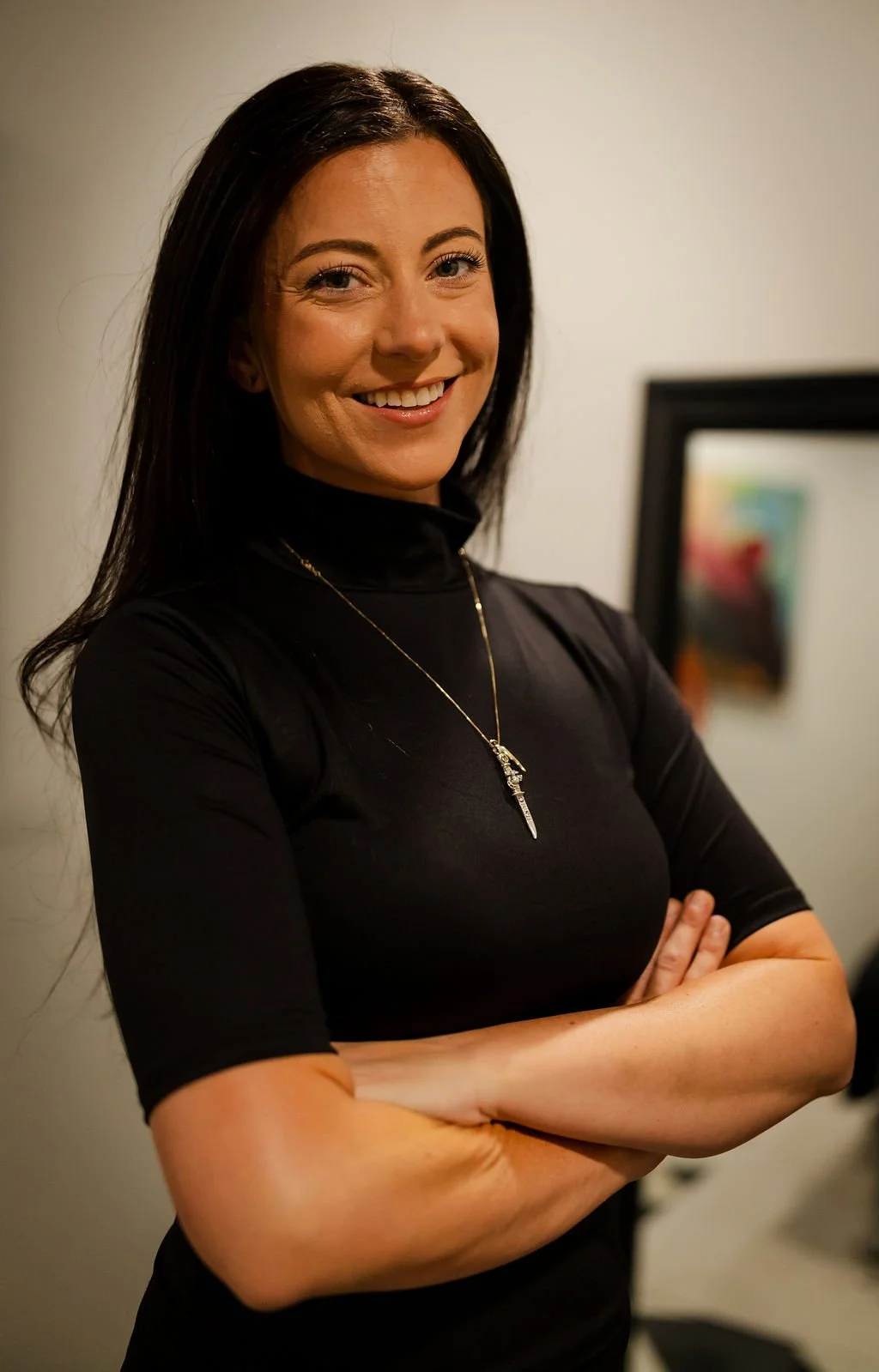 A woman with long dark hair smiling and crossing her arms, wearing a black top and a pendant necklace, standing indoors.