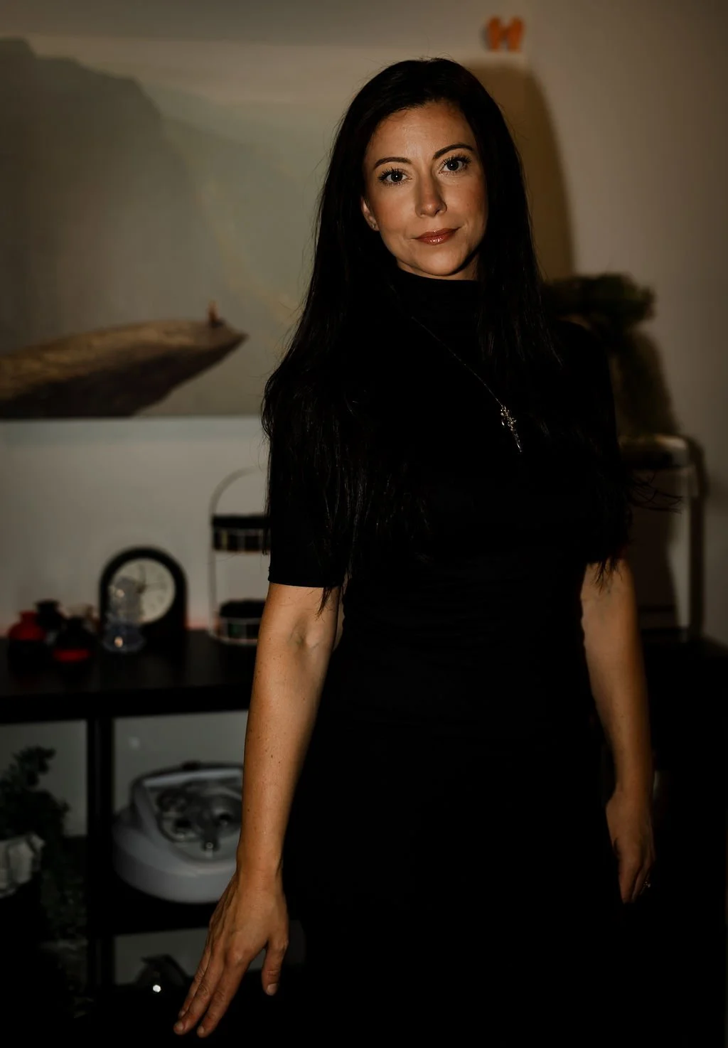 A woman with long dark hair wearing a black top standing indoors with a neutral expression, in front of a dark background with a shelf containing various decorative items.