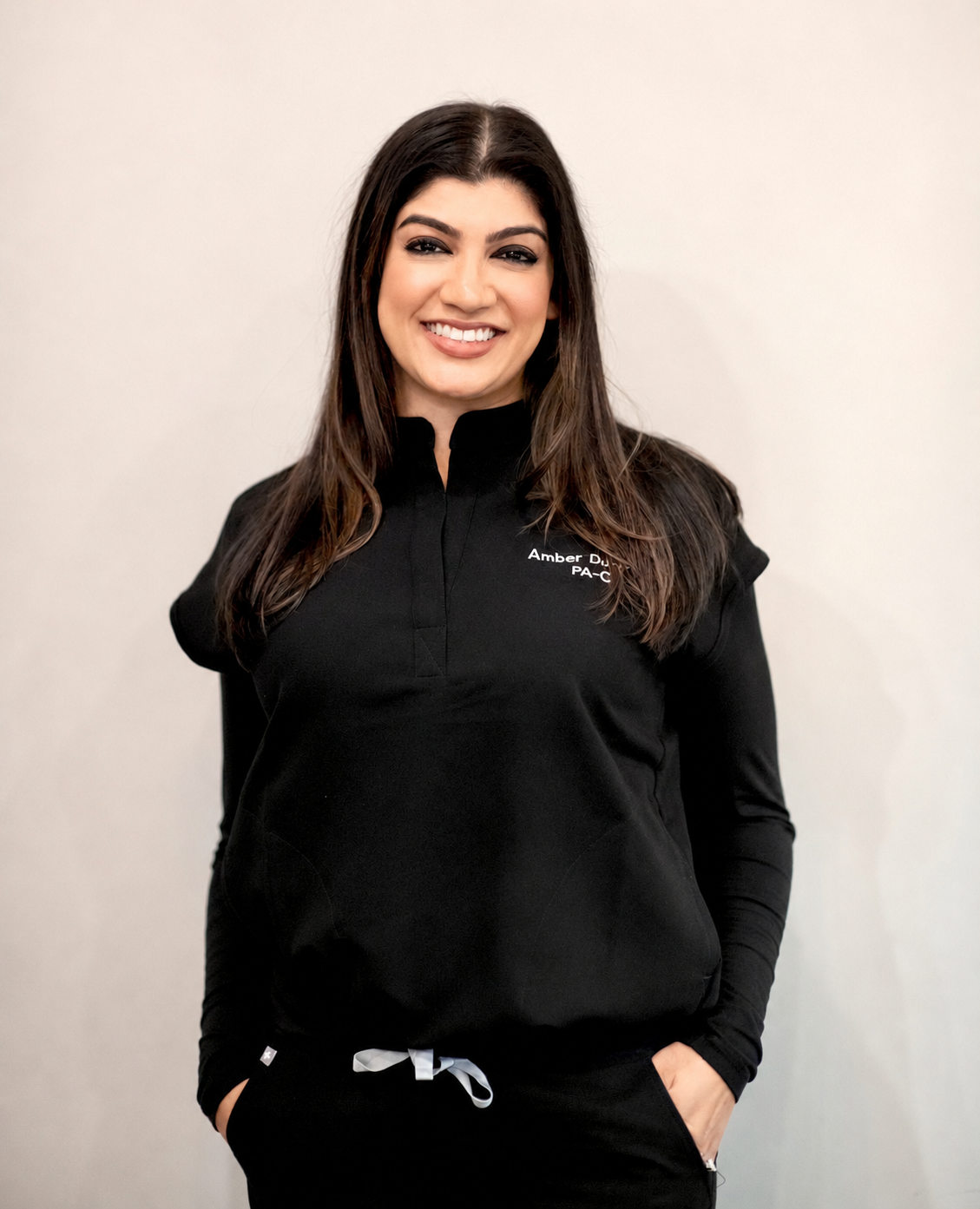 A woman with long brown hair smiling and wearing a black quarter-zip shirt with white text that says "Amber D... PA-" standing against a plain off-white background.