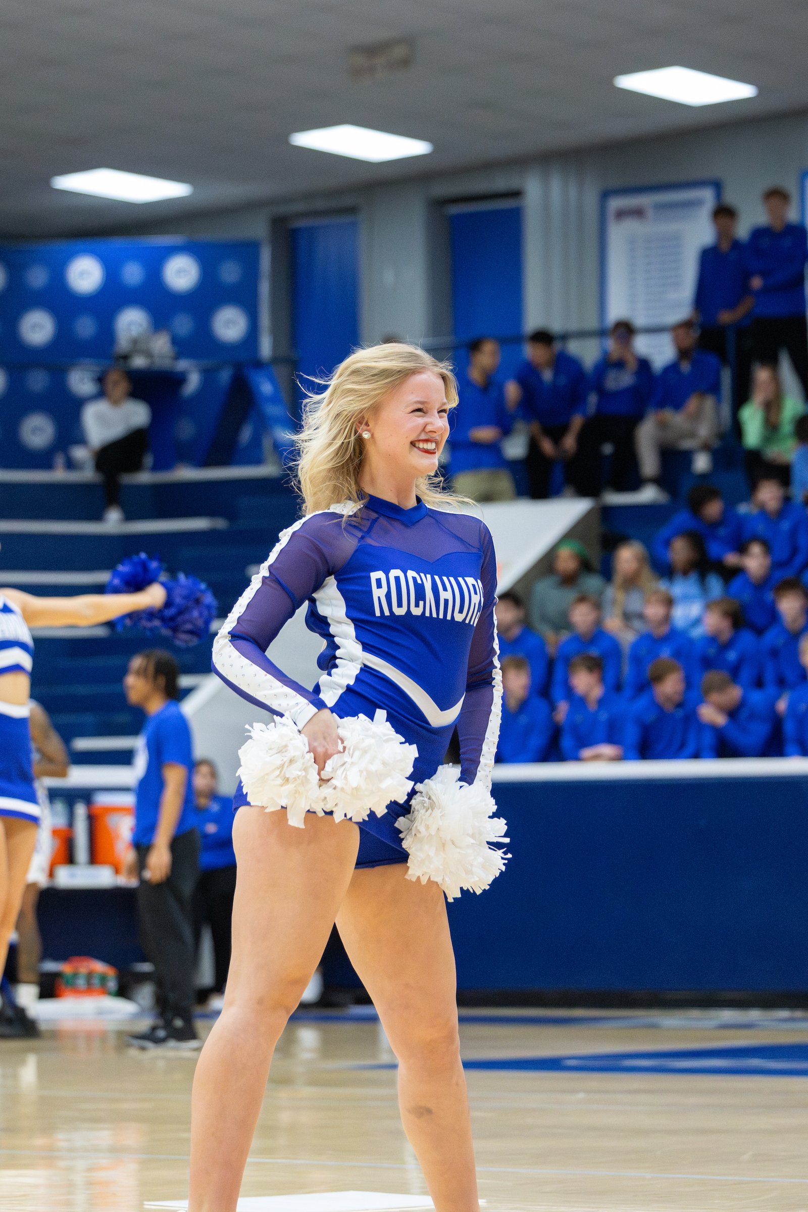 Cheerleader in blue and white uniform holding white pom-poms in a gymnasium with spectators and team members in the background.