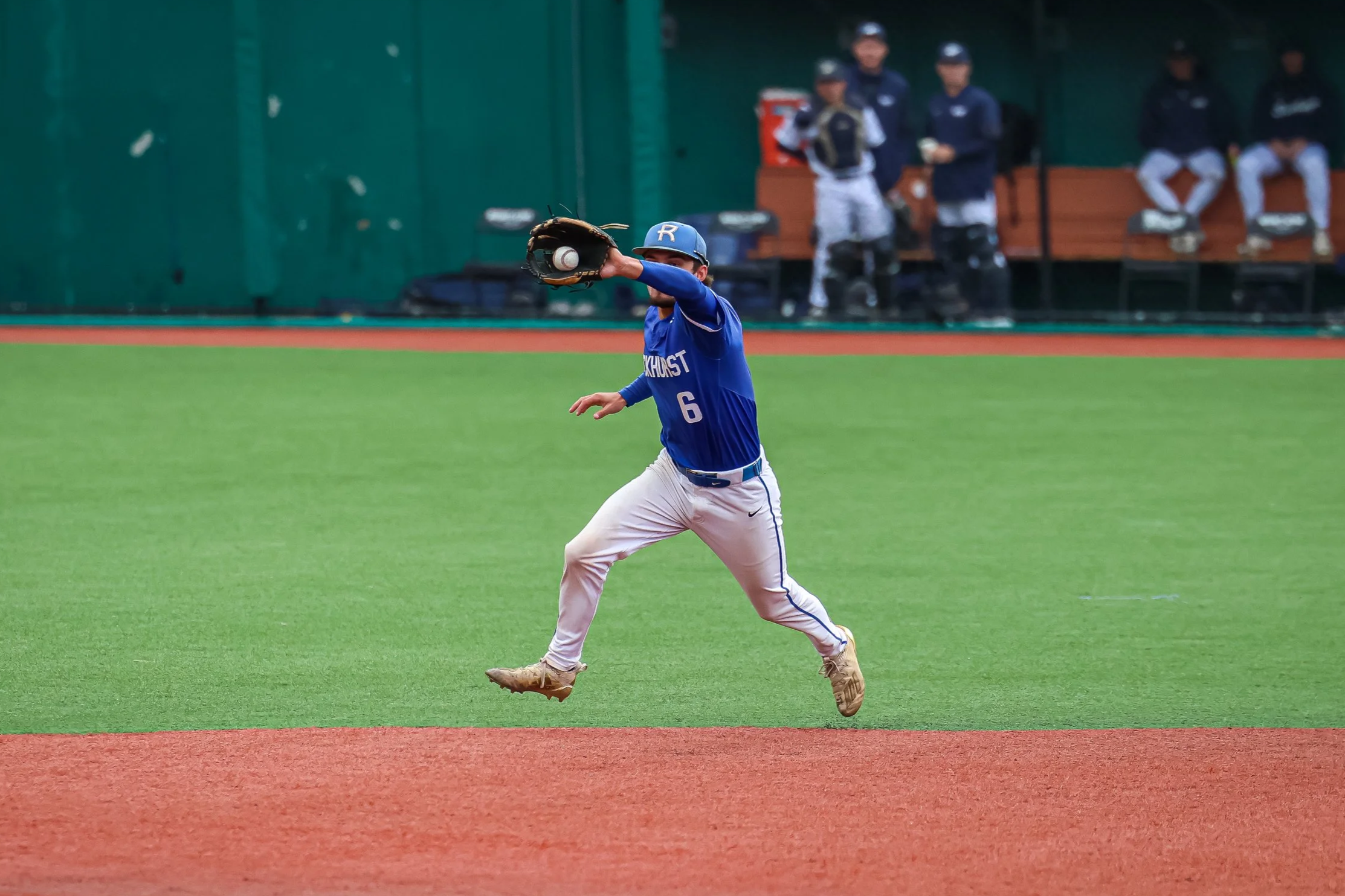 A baseball player in a blue jersey and white pants fielding a ball on a baseball field, with other players sitting on the bench in the background.