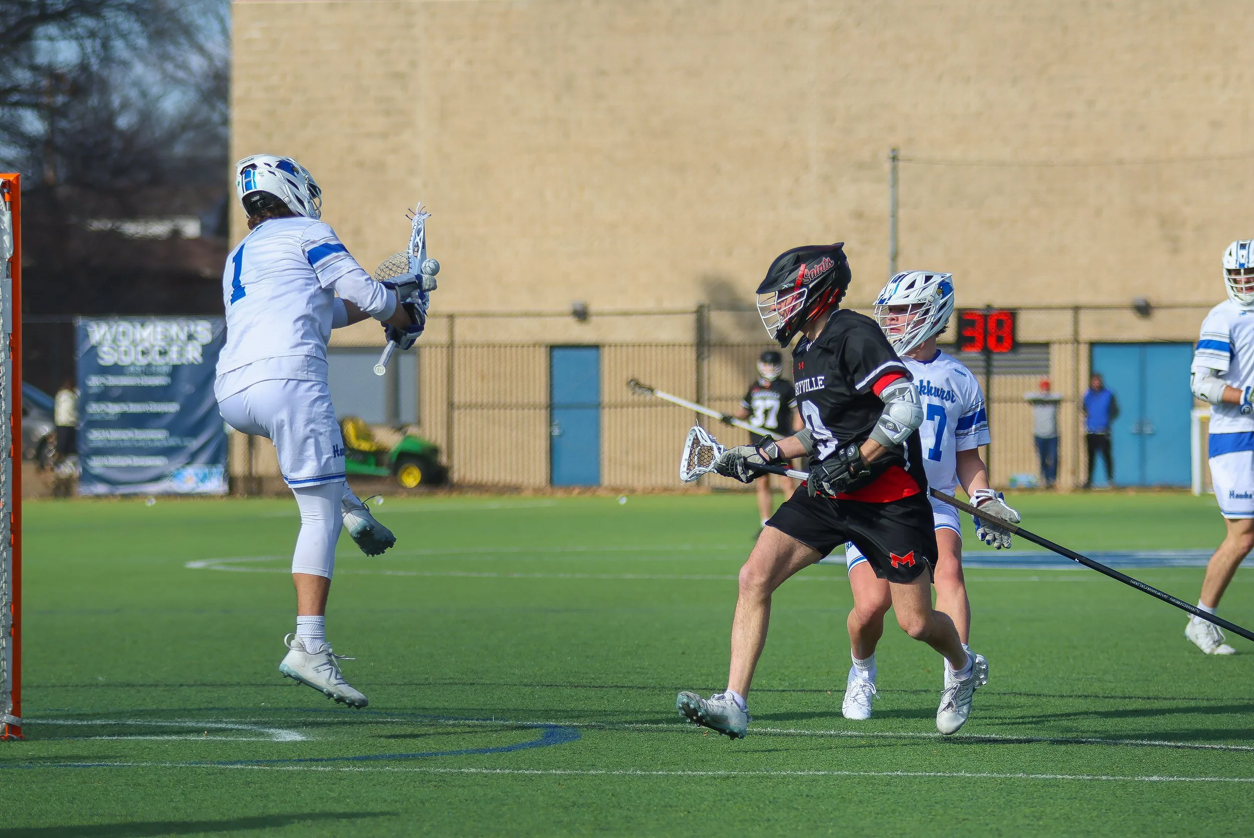 Lacrosse players on a green field during a game, with one player jumping to catch the ball in mid-air, and others running with lacrosse sticks. A large beige wall and scoreboard are in the background.