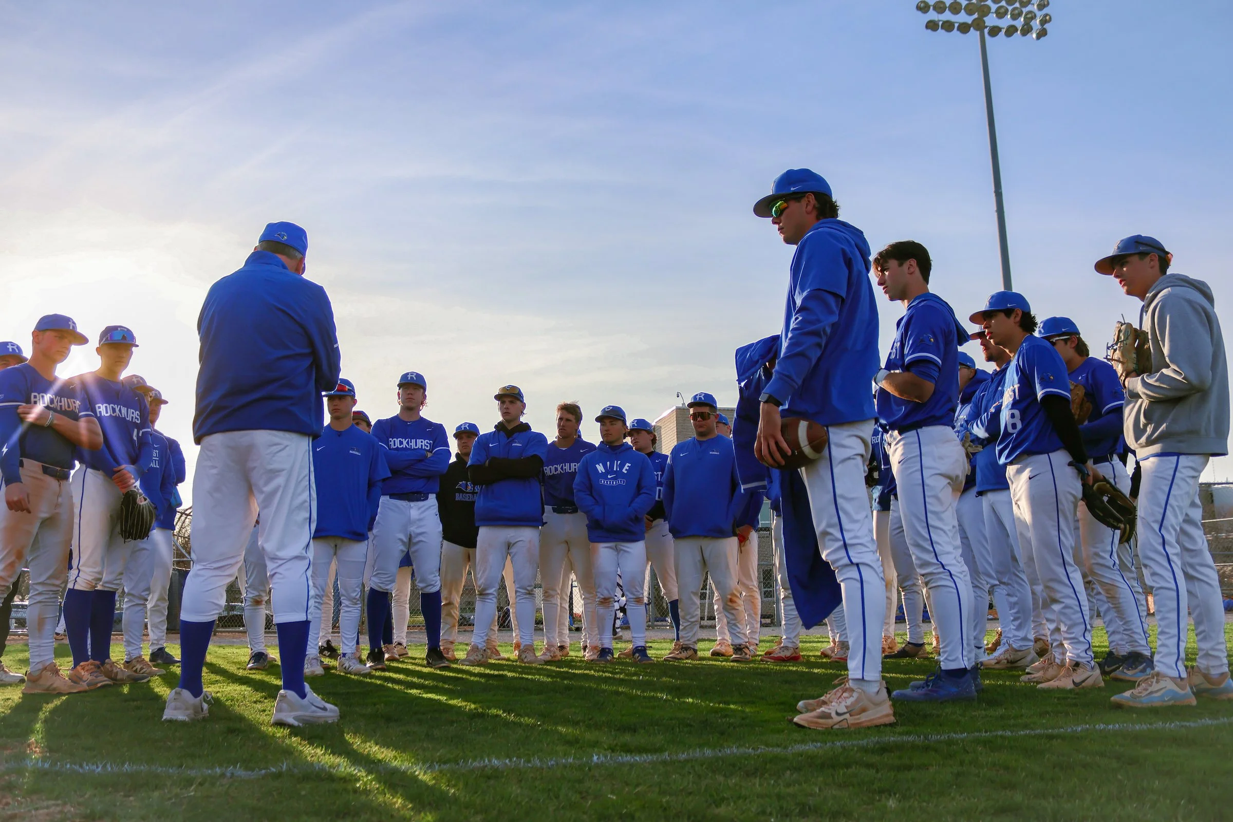 Youth baseball team in blue uniforms gathered around coach on field, possibly during a game or practice, with sunlight and blue sky overhead.