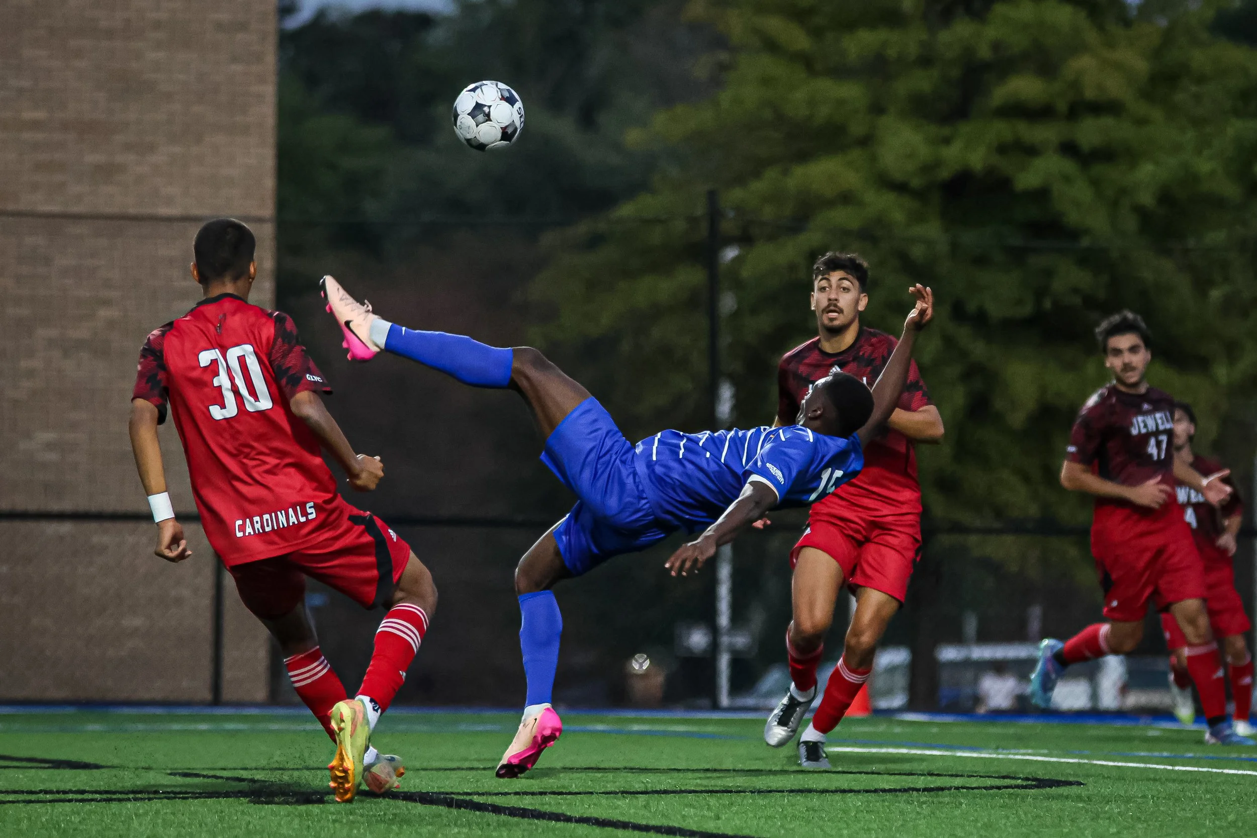 Soccer players in action during a game, with a player in blue executing a high kick to reach the ball amidst opponents in red jerseys.