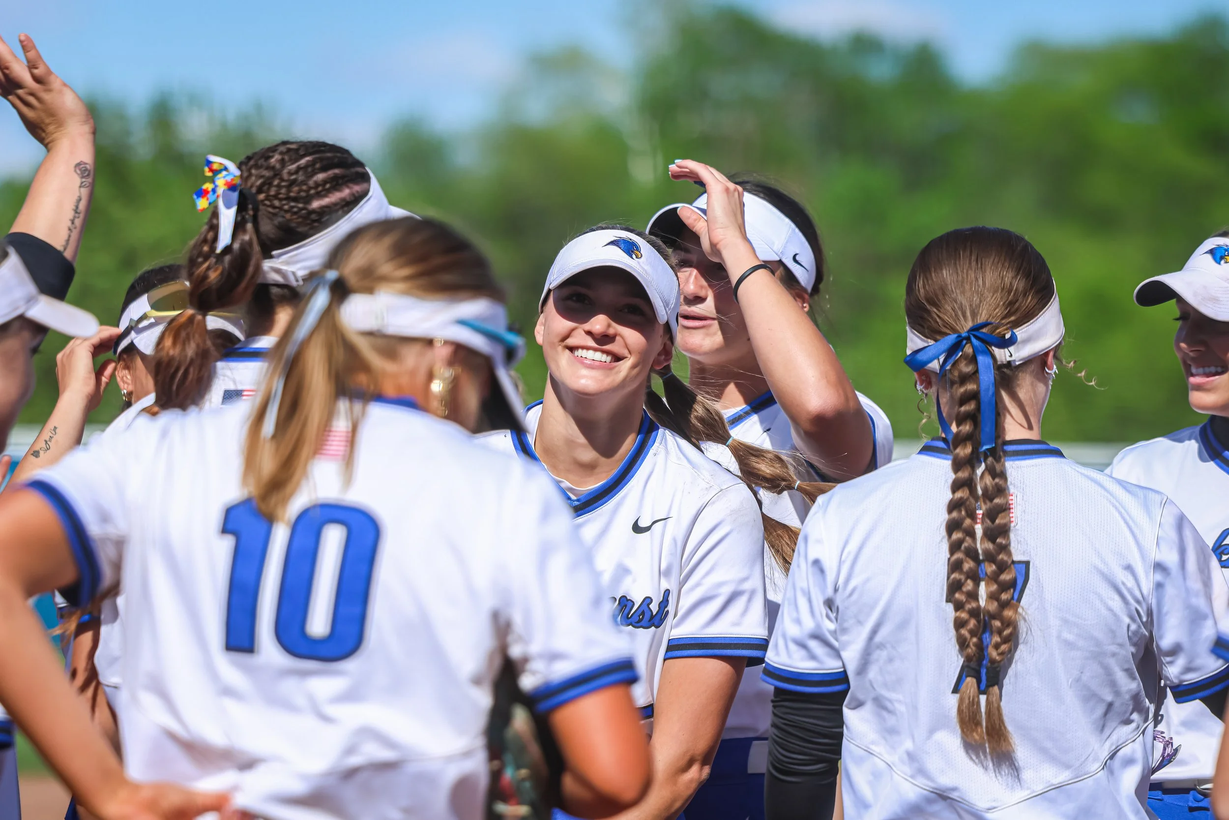 A group of female softball players wearing white uniforms with blue accents huddled together during a game.
